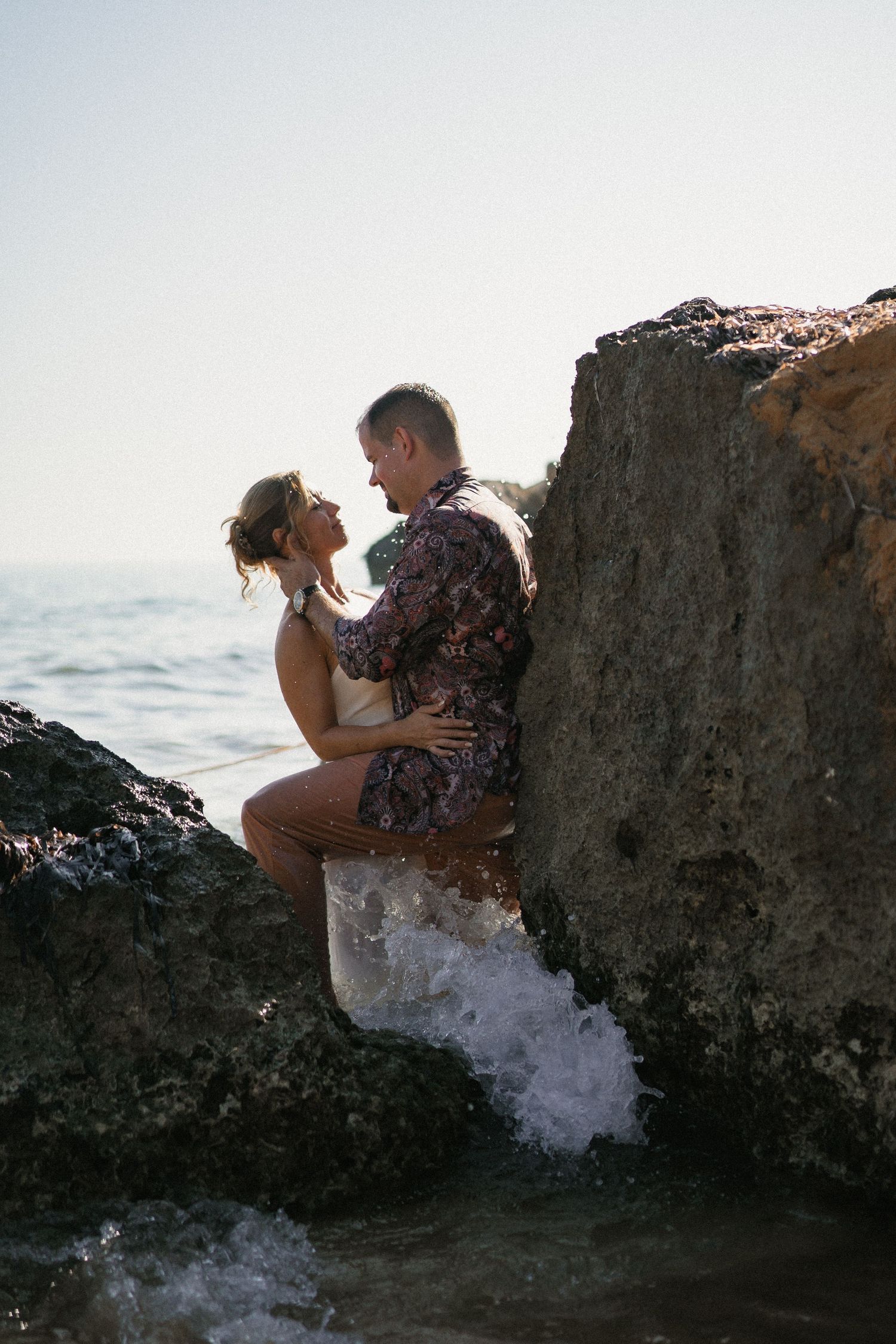 A couple embraces against dark coastal rocks as waves splash around them at the beach.