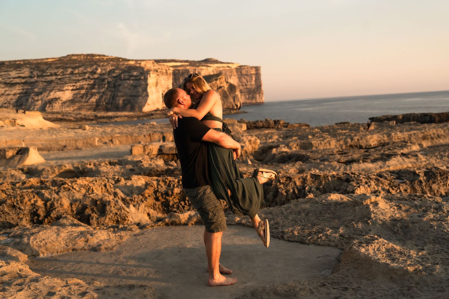 Couple embracing at golden hour on rocky coastal cliffs with ocean views in warm sunset light.