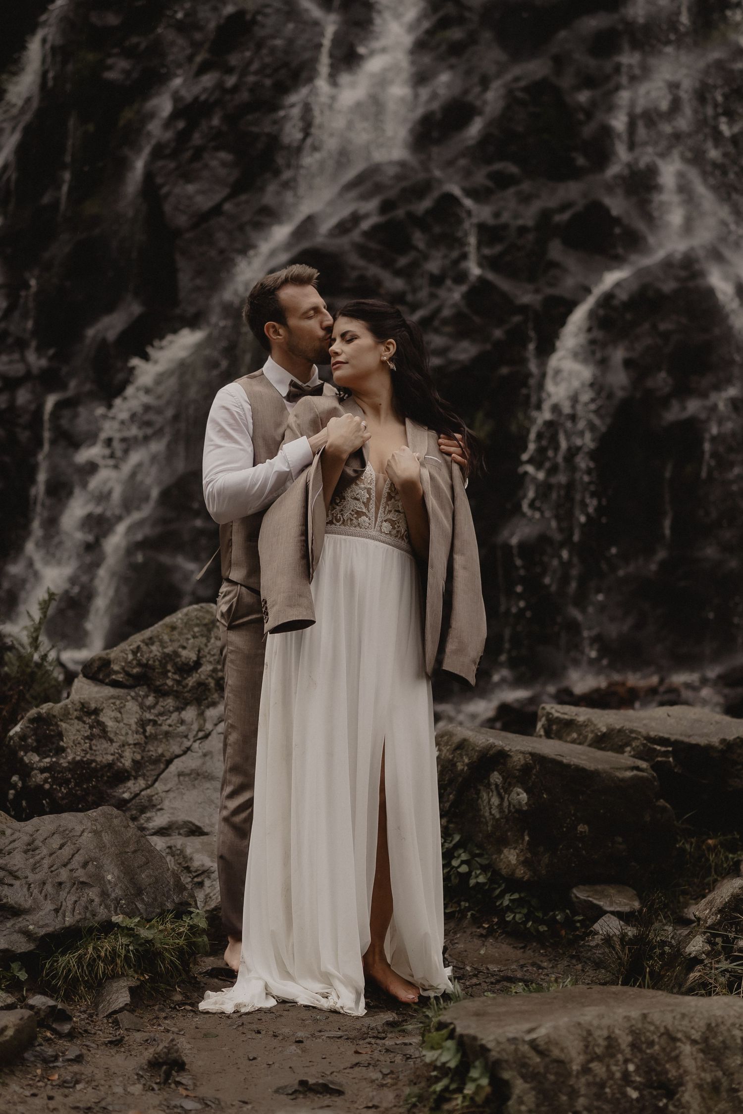 Bride and groom embrace before a waterfall, wearing elegant wedding attire in a romantic outdoor ceremony.