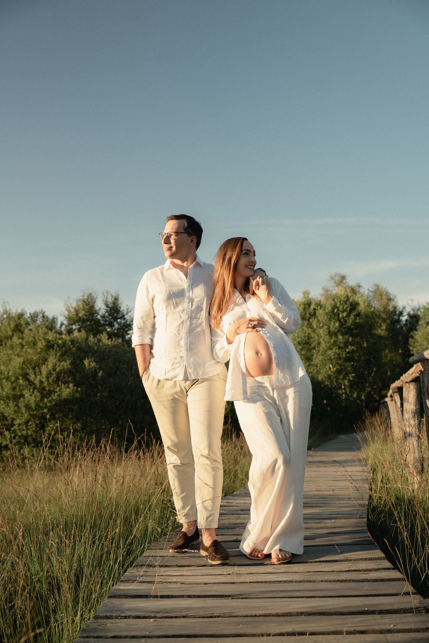 Pregnant couple in white outfits walking on a wooden boardwalk surrounded by green trees under a clear blue sky.