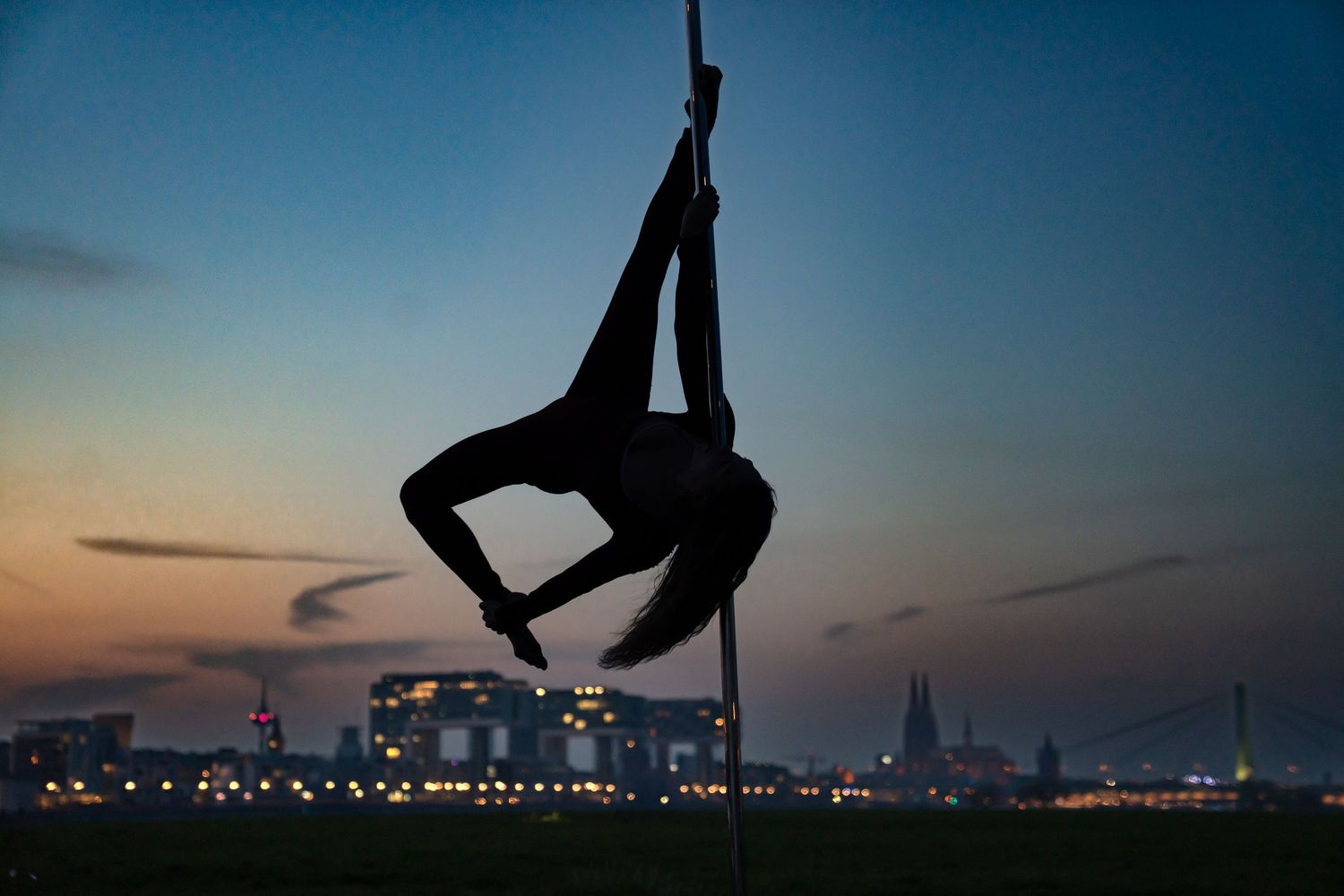 Silhouette of a pole dancer performing an aerial split against a stunning city skyline at dusk.