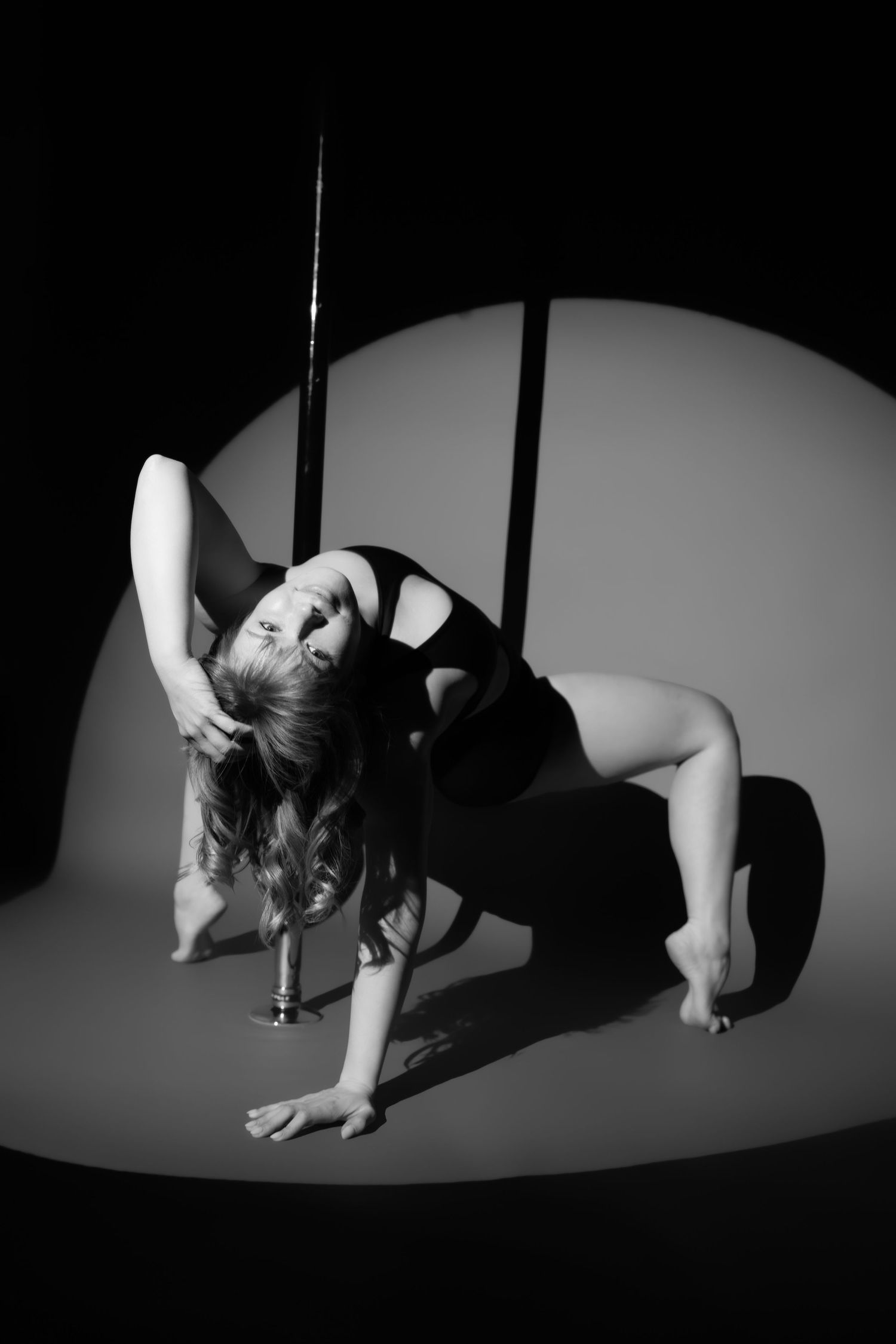 Black and white photo of a flexible woman posing gracefully beside a pole in dramatic spotlight lighting.