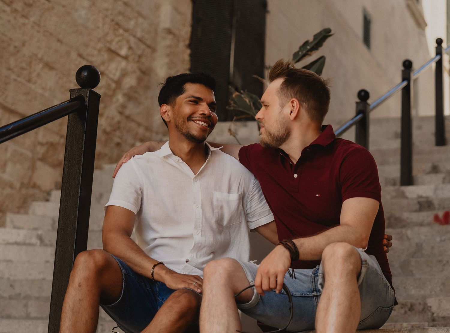 Two men sitting on outdoor stone steps, smiling and looking at each other in a sunny Mediterranean setting.