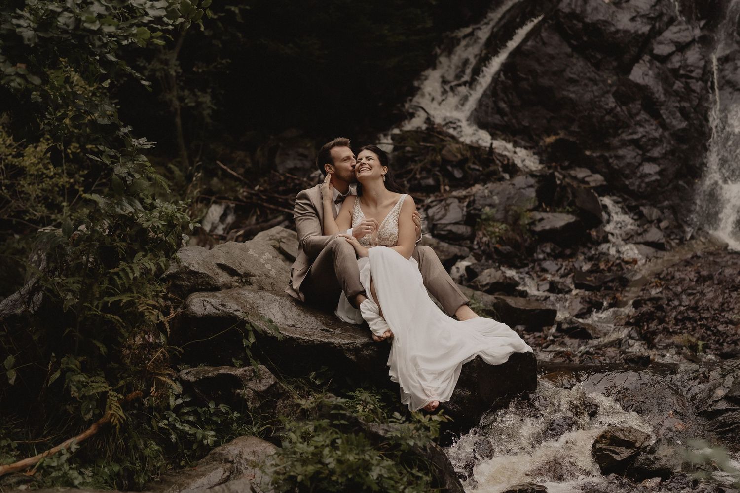 Newlywed couple sits together on rocky mountain ledge overlooking scenic valley during outdoor wedding photoshoot.