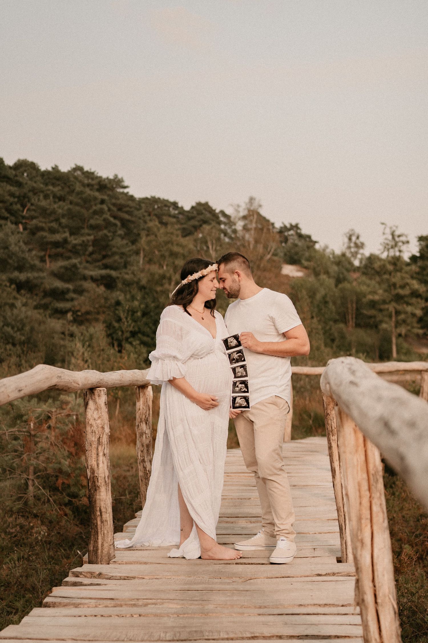 A couple sits together on a wooden boardwalk holding ultrasound photos during a maternity photoshoot.