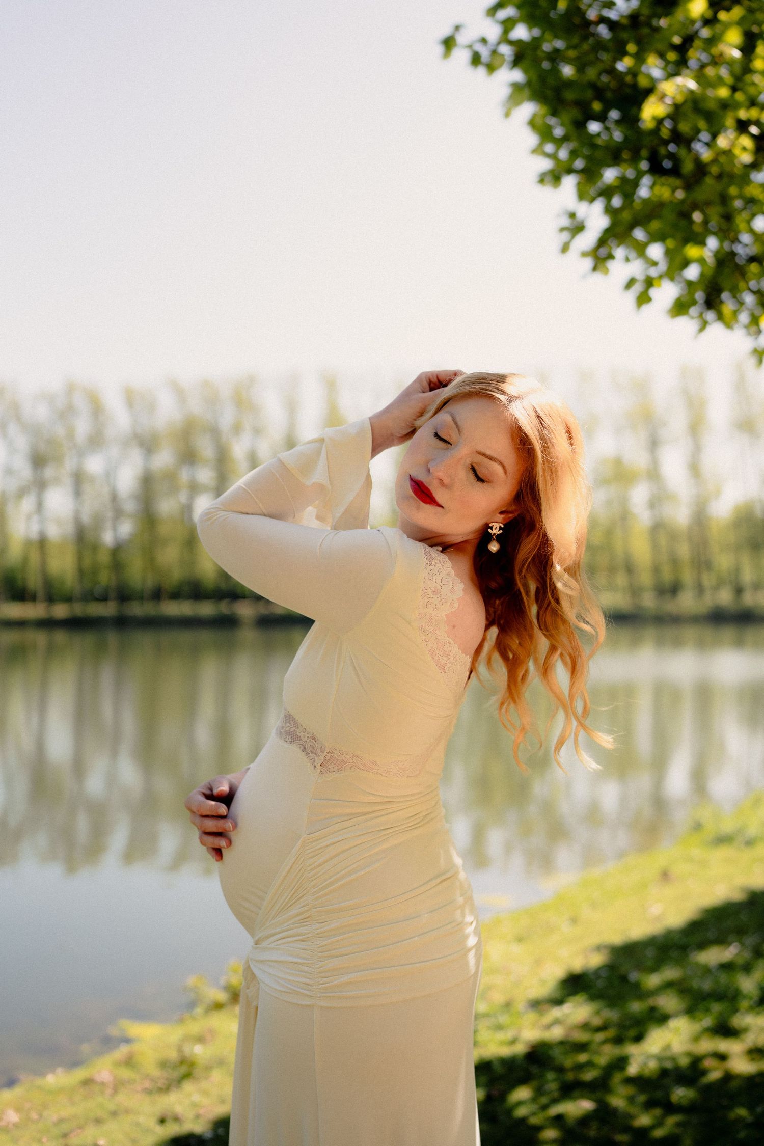 Elegant pregnant woman in white gown posing gracefully beside a serene lake at golden hour.