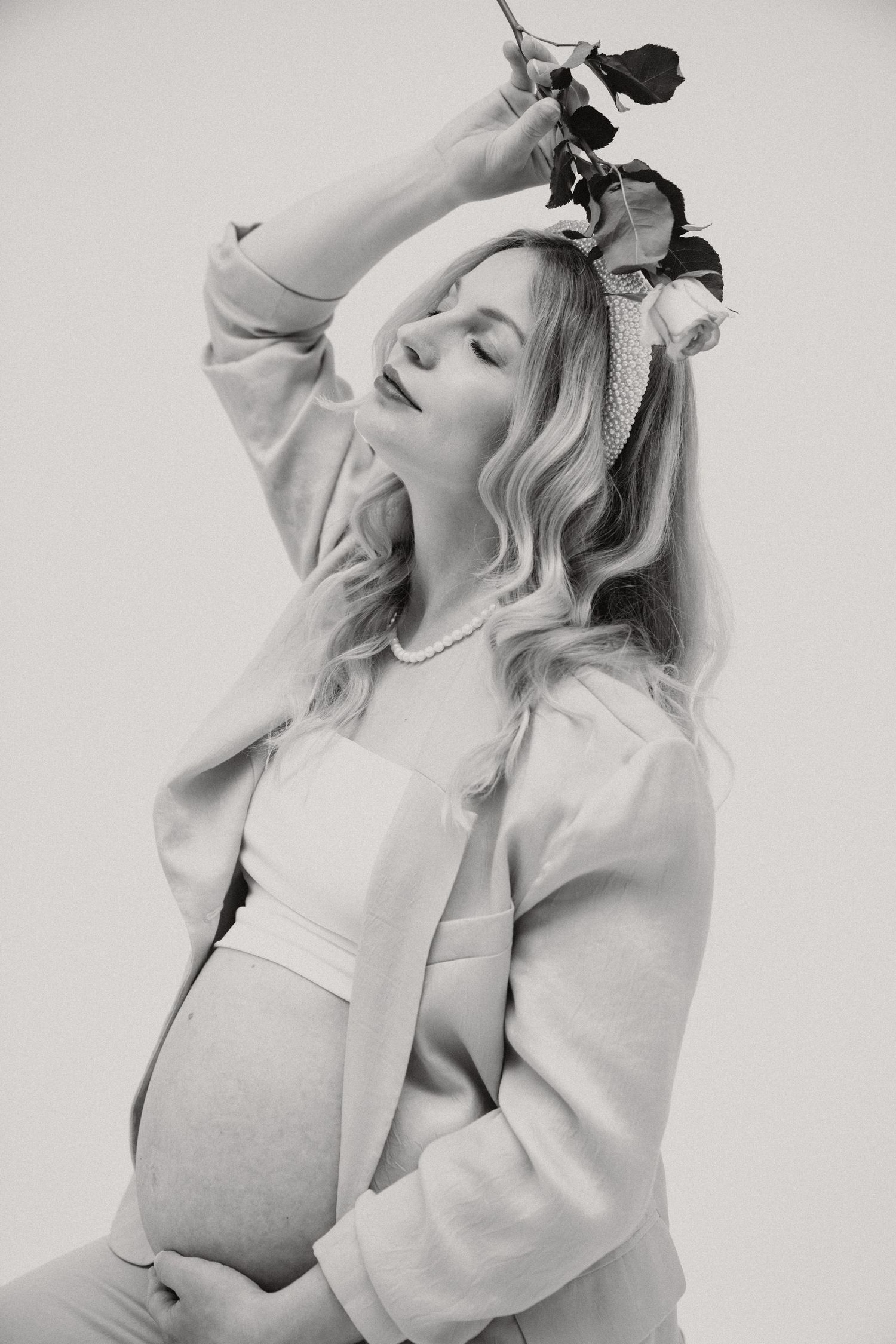 Black and white photo of a pregnant woman holding a rose above her head, wearing a casual outfit.