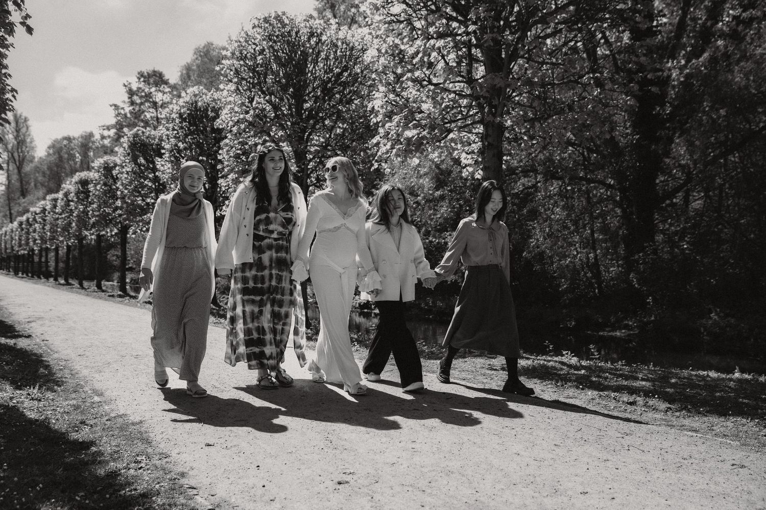 Group of five women walking together on a tree-lined path in a black and white outdoor photo.