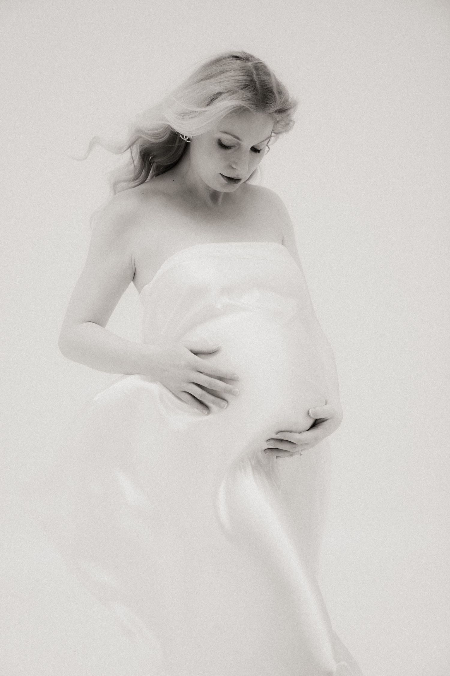 Black and white maternity photo of a blonde woman in white fabric holding her pregnant belly.