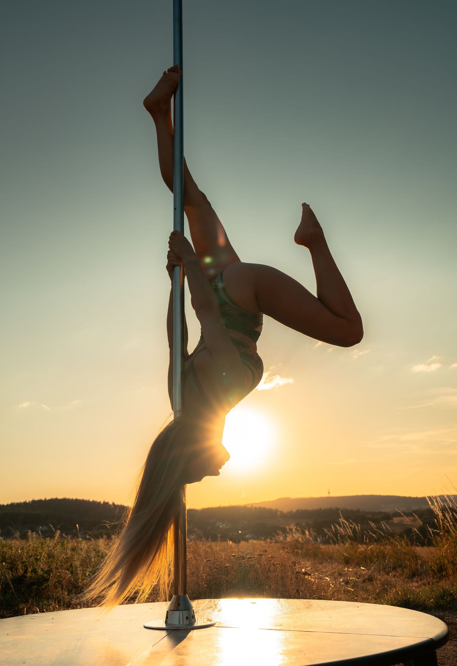 Silhouette of a woman performing pole dancing outdoors at golden sunset with long hair flowing and legs extended gracefully.