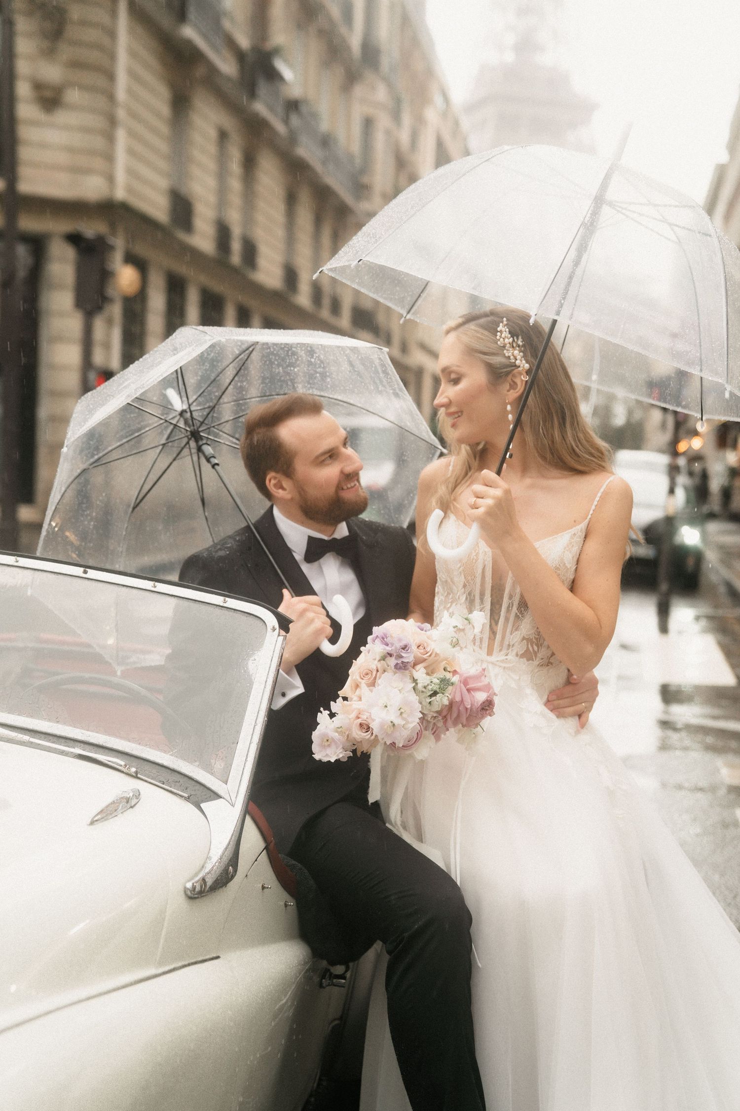 A romantic wedding couple shares intimate moments under clear umbrellas on a rainy city street in Paris.