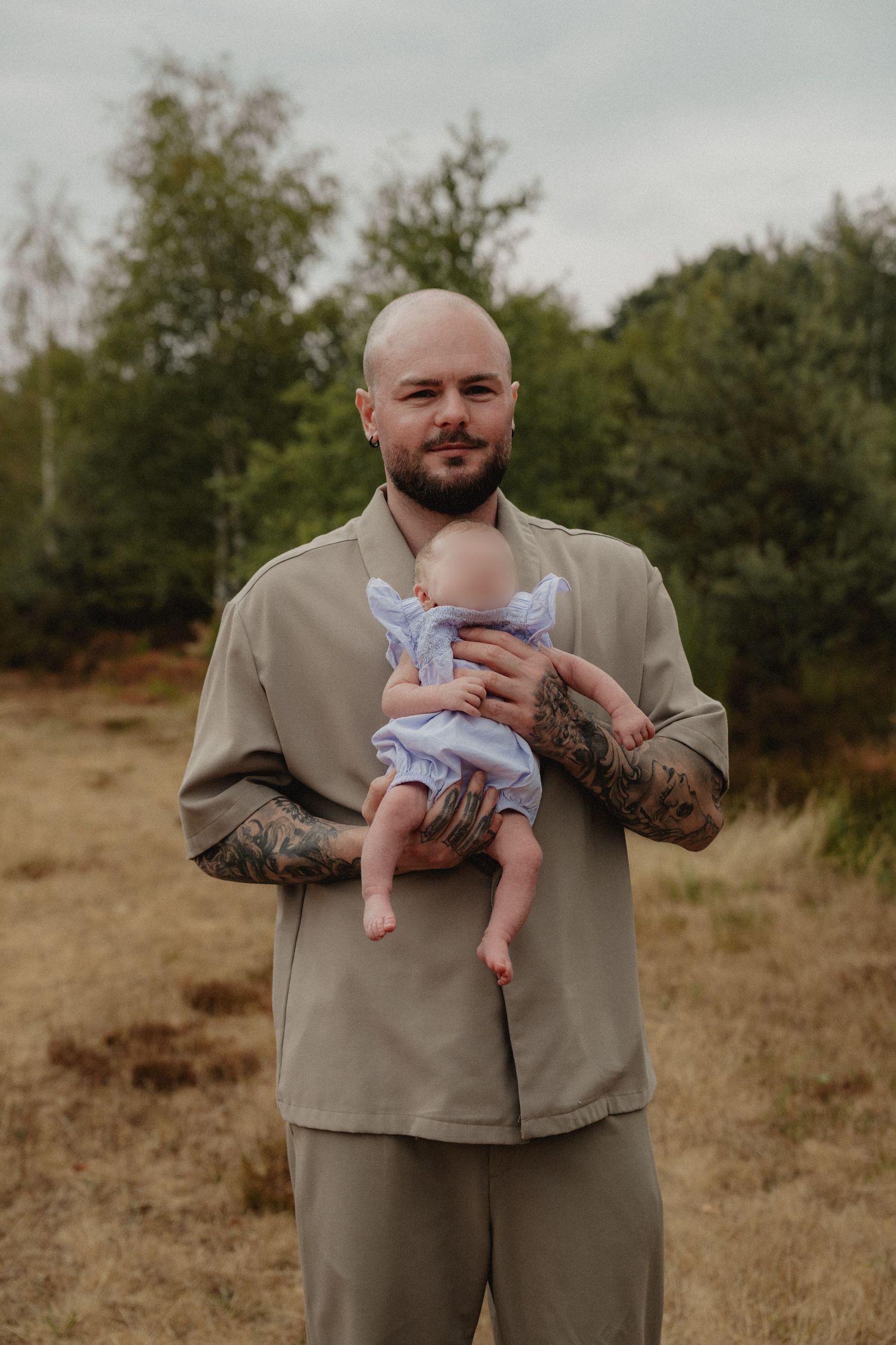 Tattooed bald man holding a baby in a purple dress outdoors in a field with green trees in background.