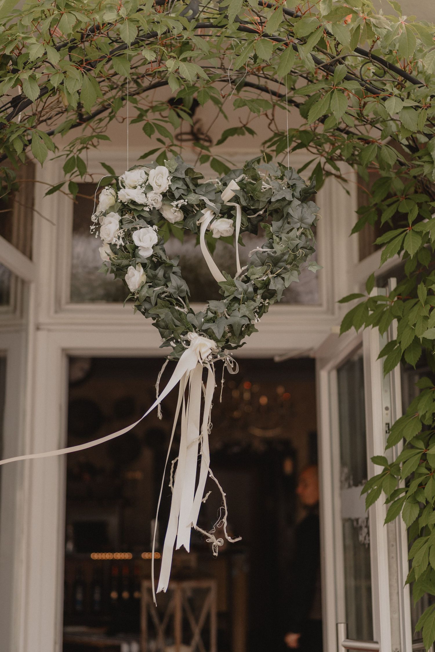 A decorative white heart-shaped wreath with floral accents hanging against a window with green foliage.