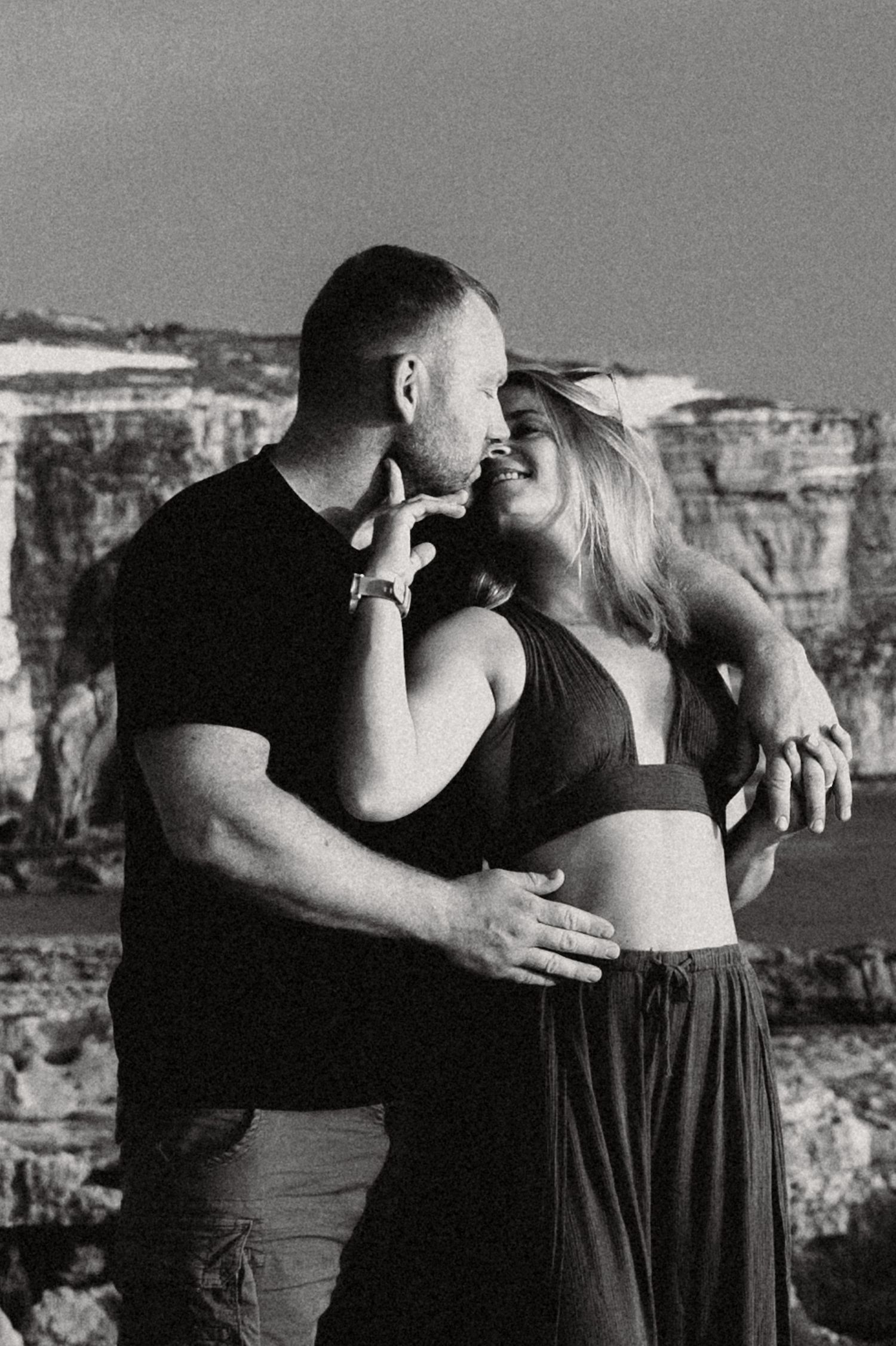 Black and white photo of a romantic couple embracing near coastal cliffs, woman in crop top smiling up at man.