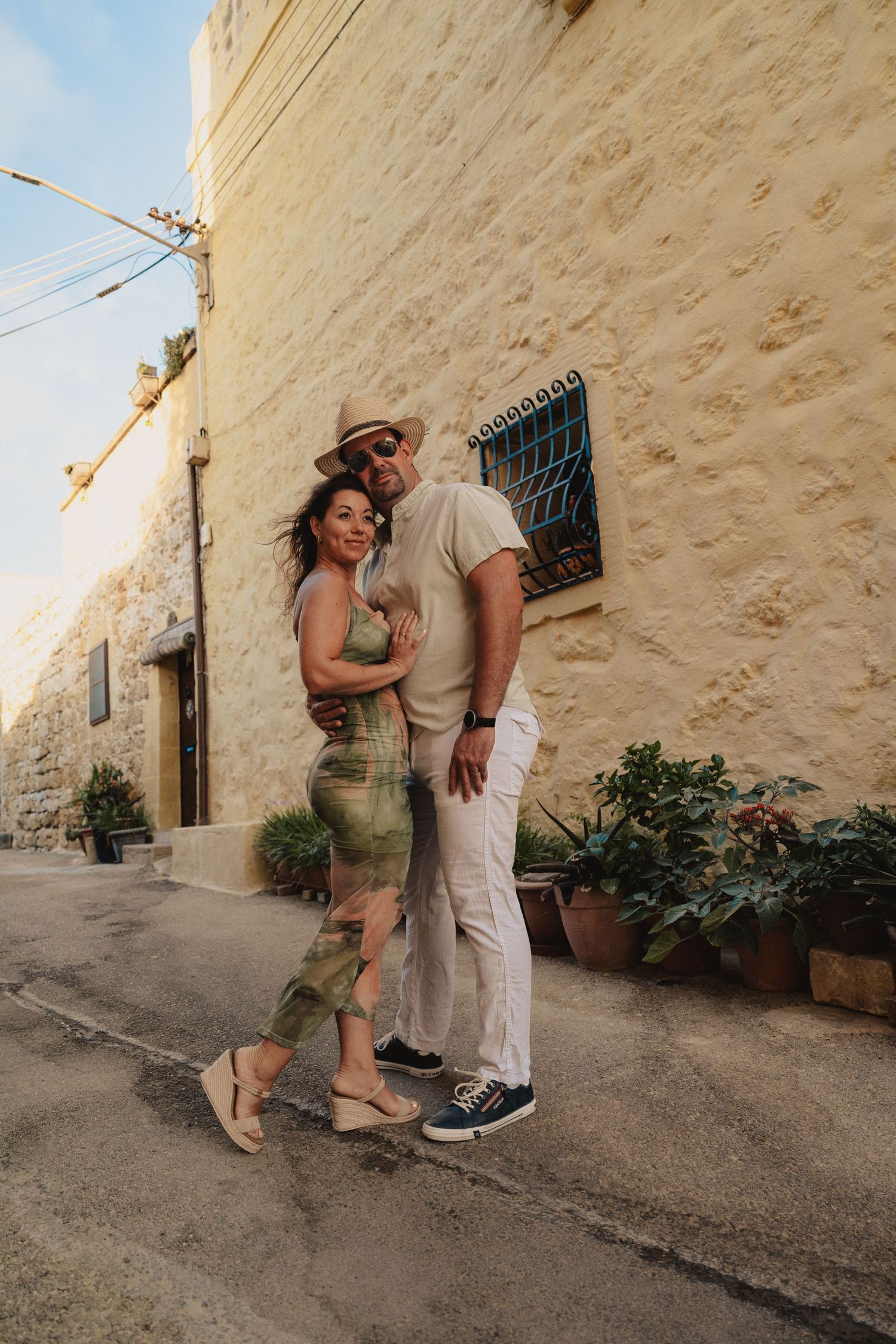Couple posing together on a sunny street beside a limestone wall with potted plants in a Mediterranean town.