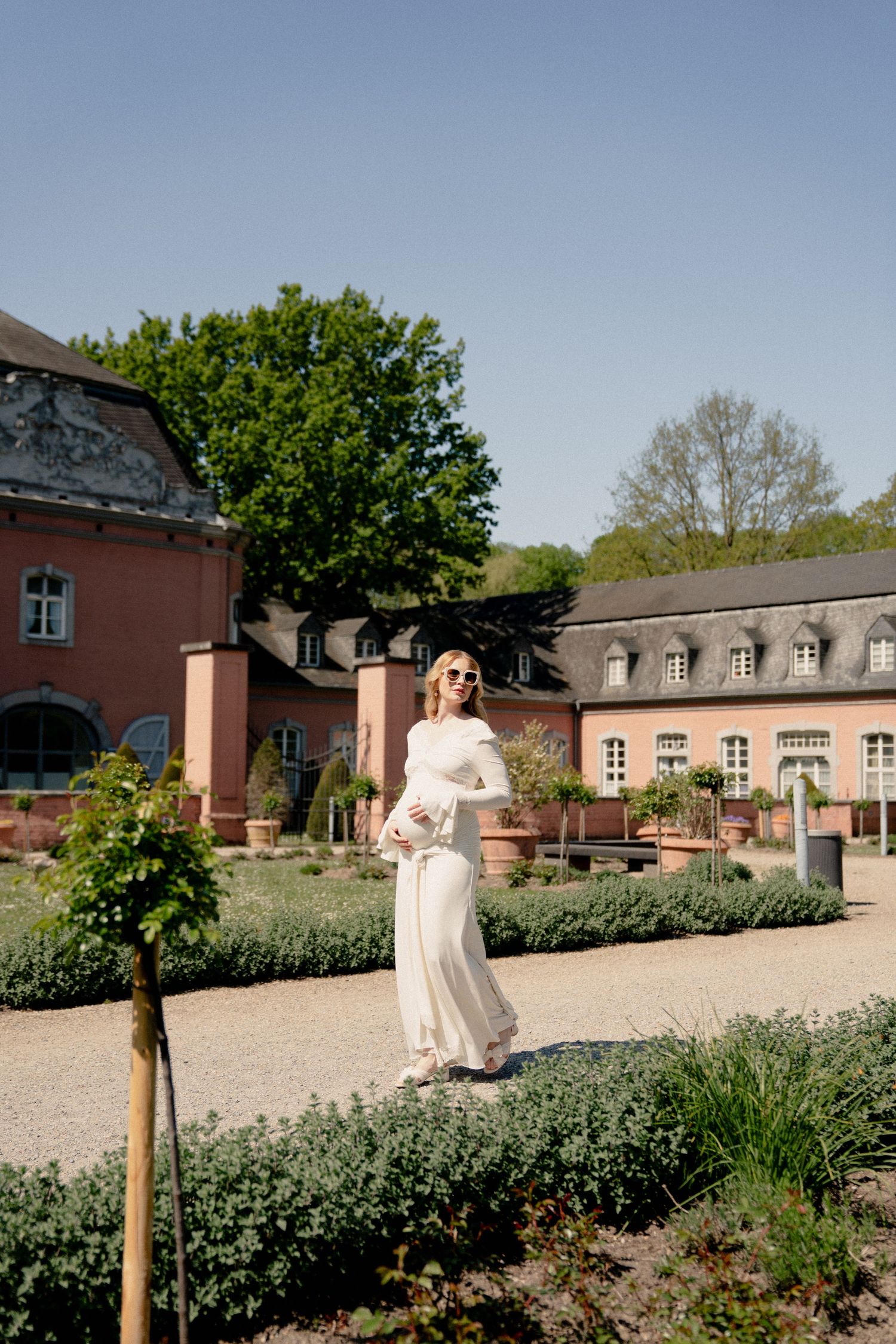 Pregnant woman in white dress standing in a formal garden courtyard of a historic estate.