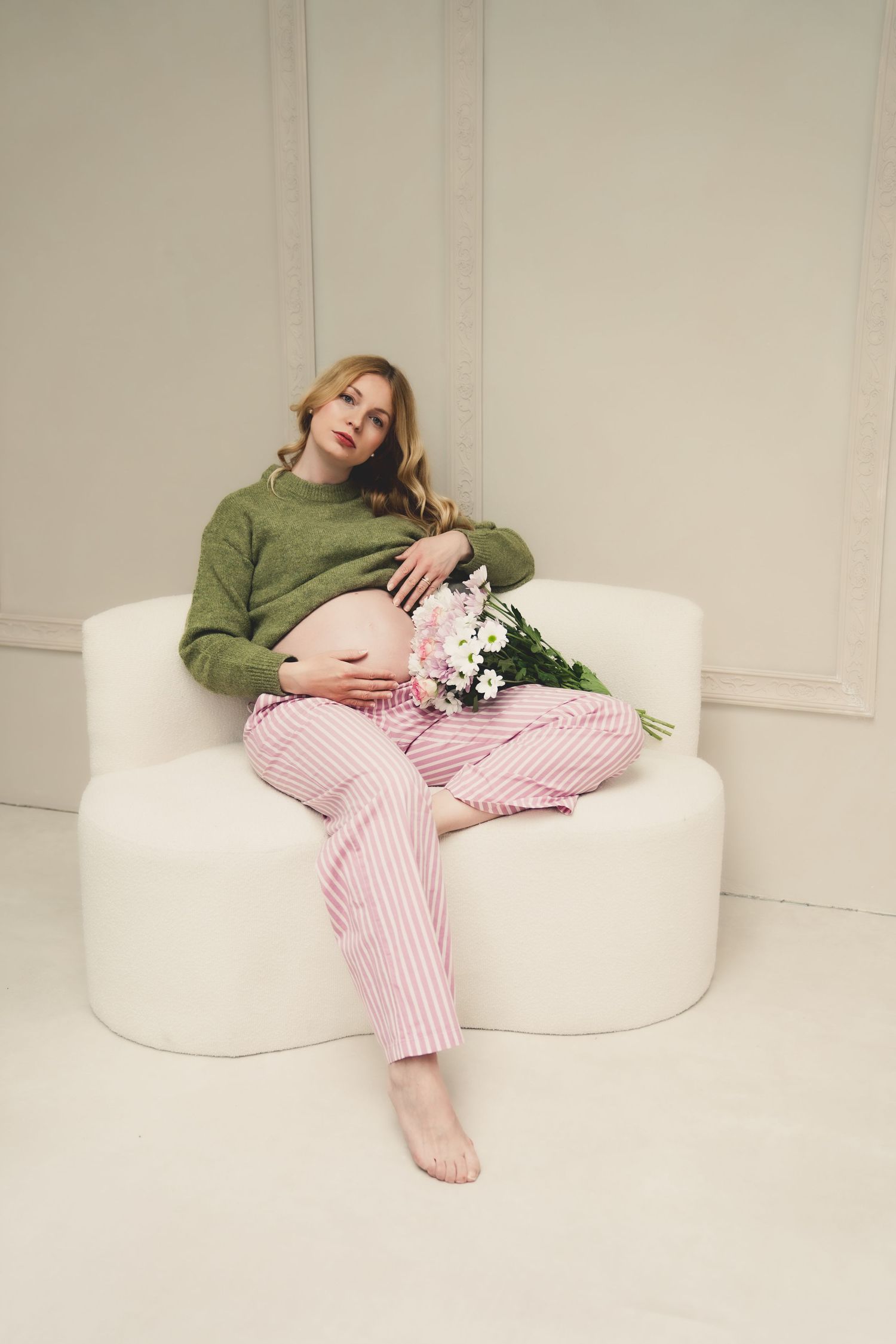 Pregnant woman in green sweater and pink striped pants sits on white chair holding flowers, cradling her baby bump.