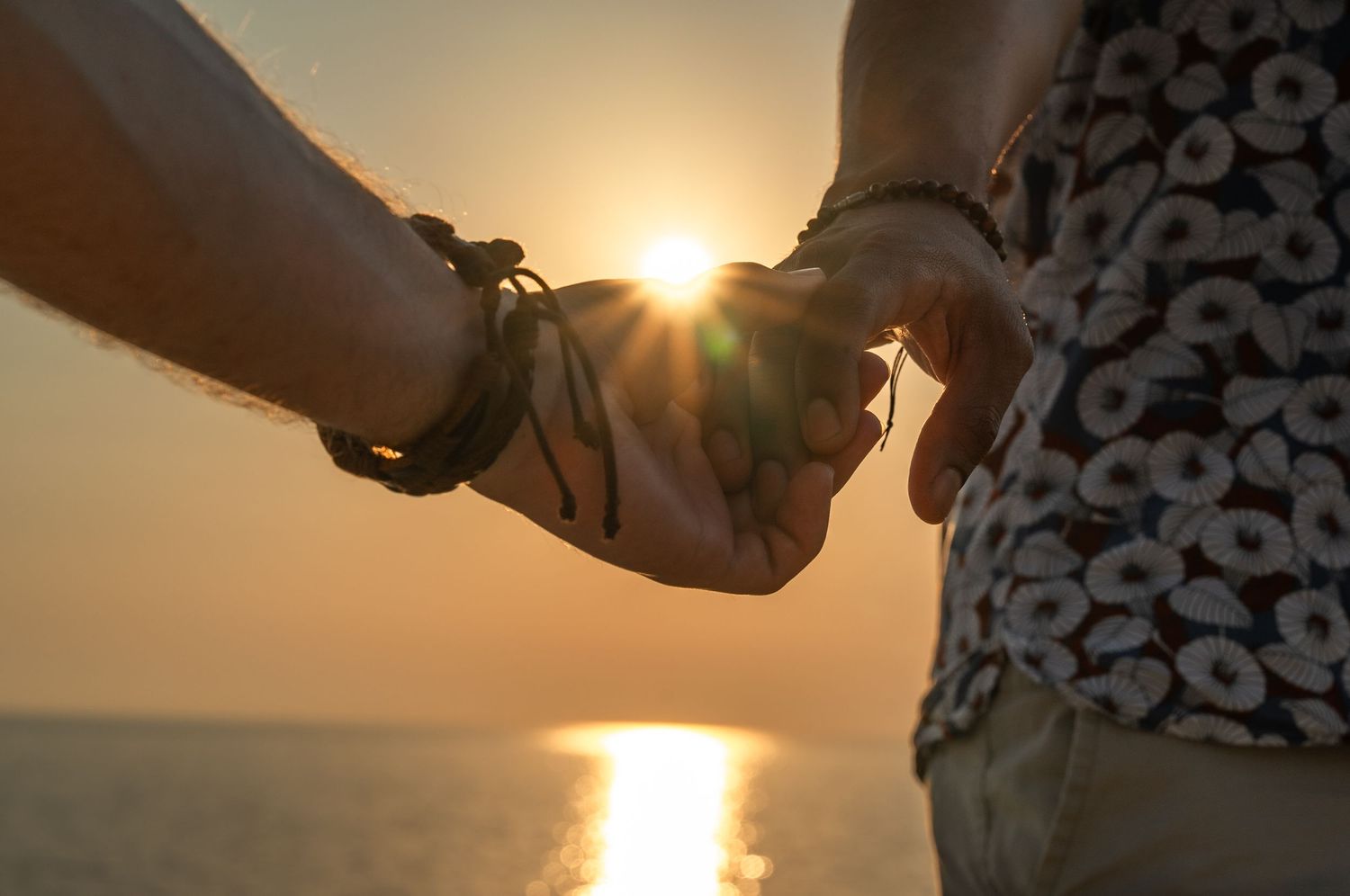 Couple holding hands at sunset by the ocean, wearing bracelets, with golden sun rays reflecting on calm water.