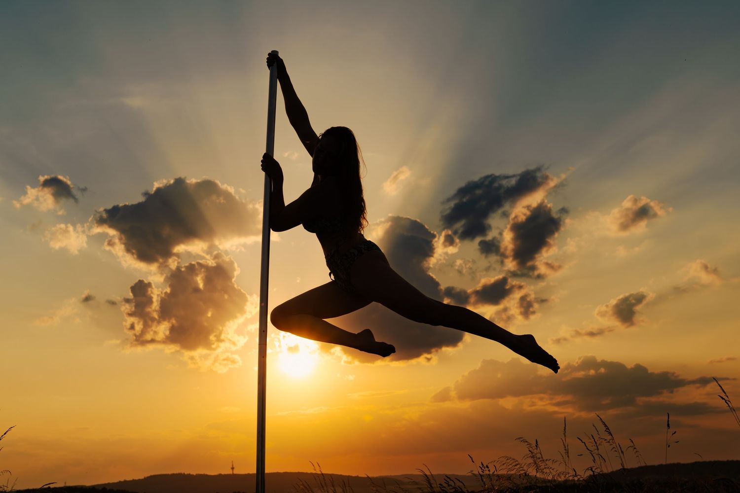 Silhouette of a woman performing pole dance against a dramatic sunset sky with golden light and clouds.