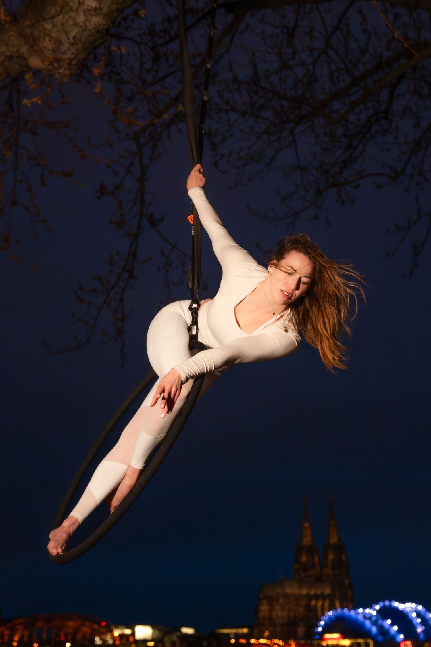 Aerial acrobat in white bodysuit performing on silk hoop suspended from tree against night city skyline.