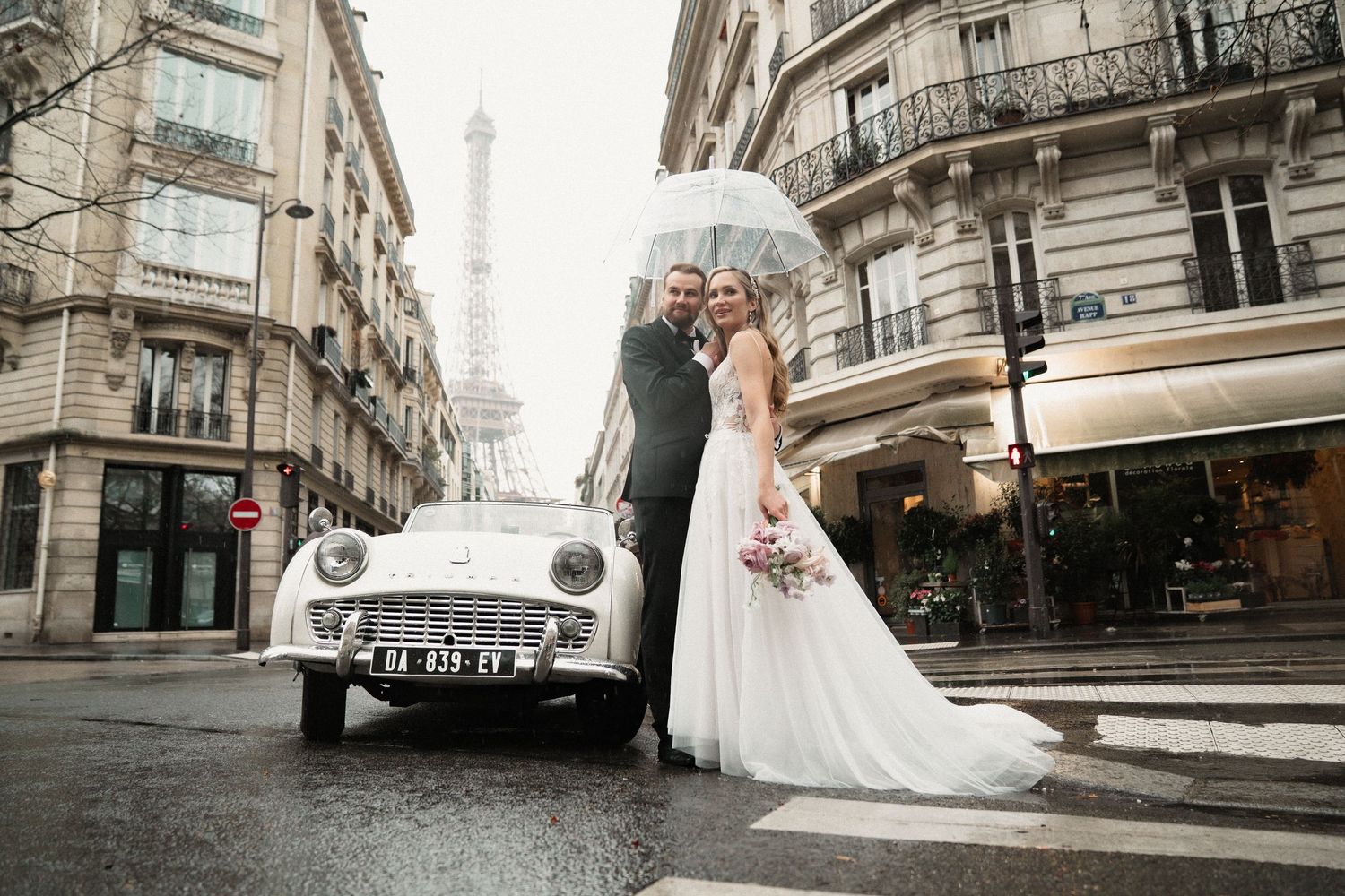 Wedding couple poses on Parisian streets with classic architecture and vintage car in background.