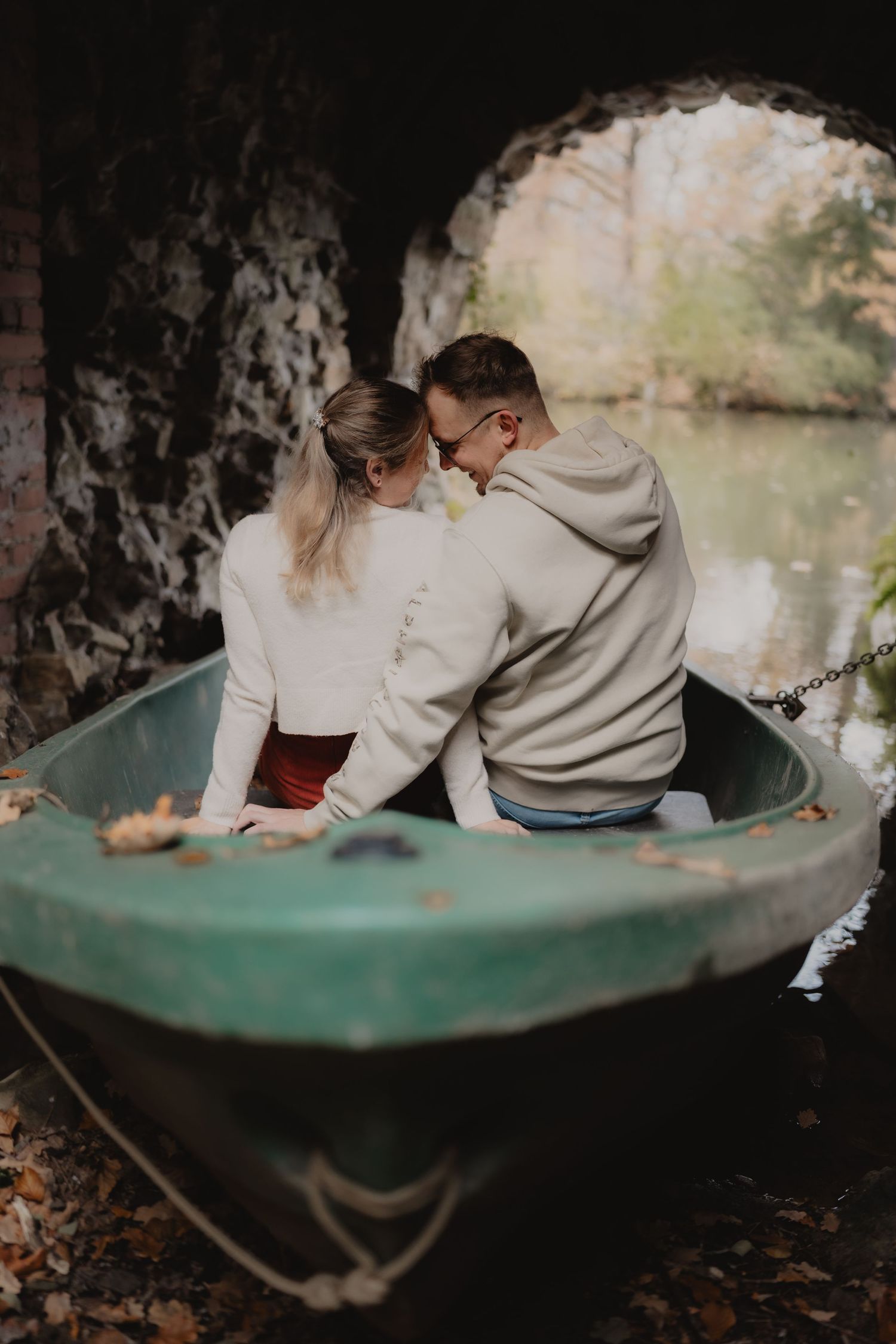 A couple sits close together on a bench near a tranquil pond on an autumn day.