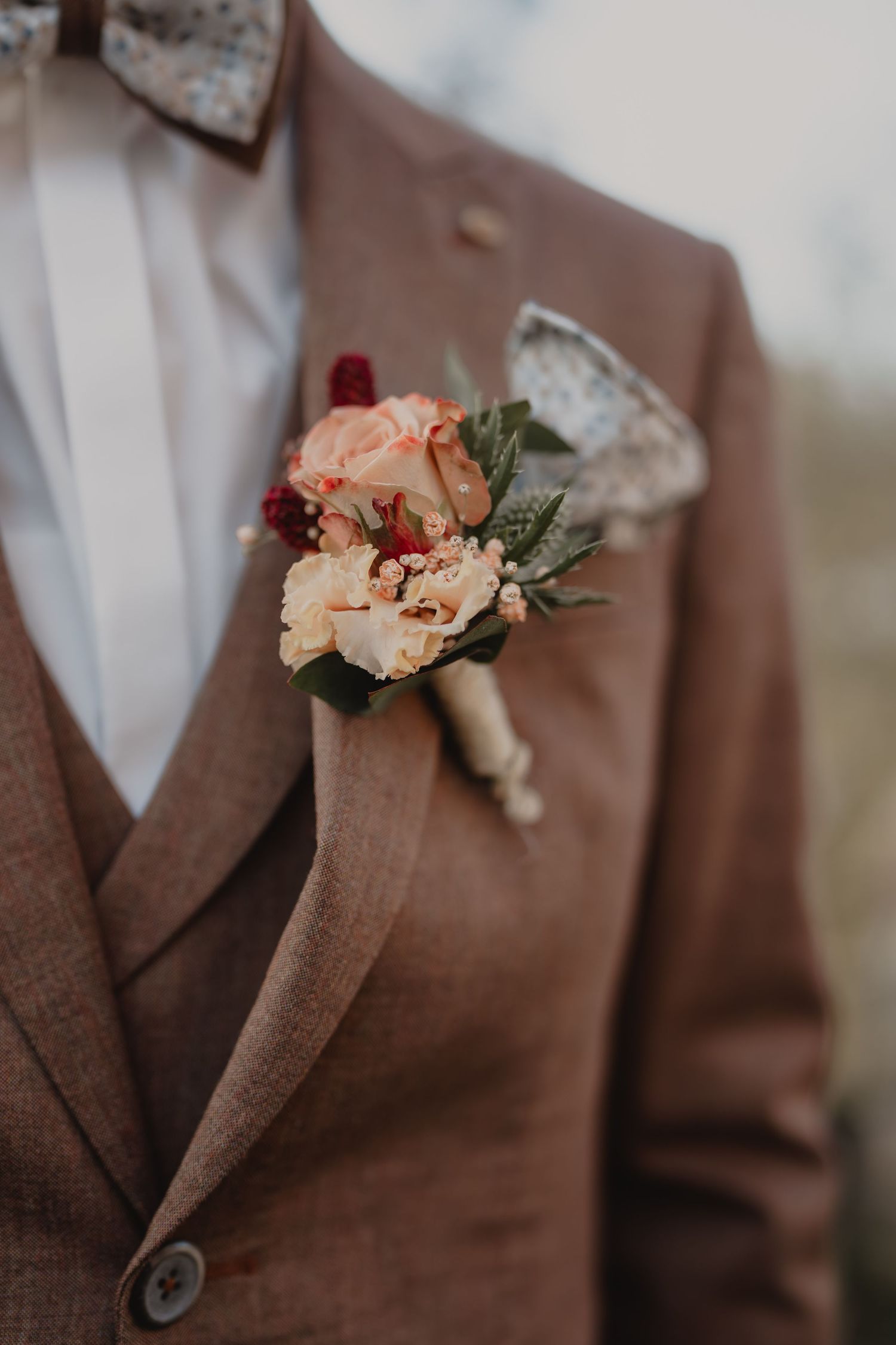 Close-up details of brown suit jackets with peach and burgundy floral boutonnieres at a formal event.