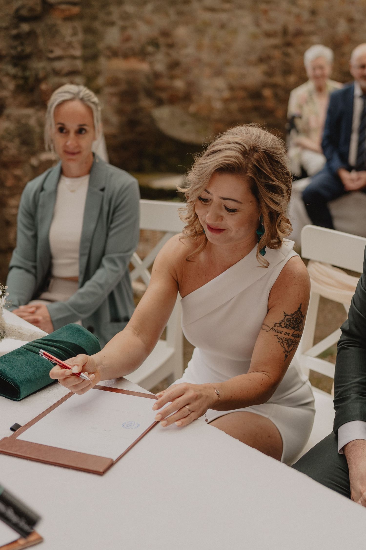 Bride in white dress signing marriage paperwork at wooden table during wedding ceremony.