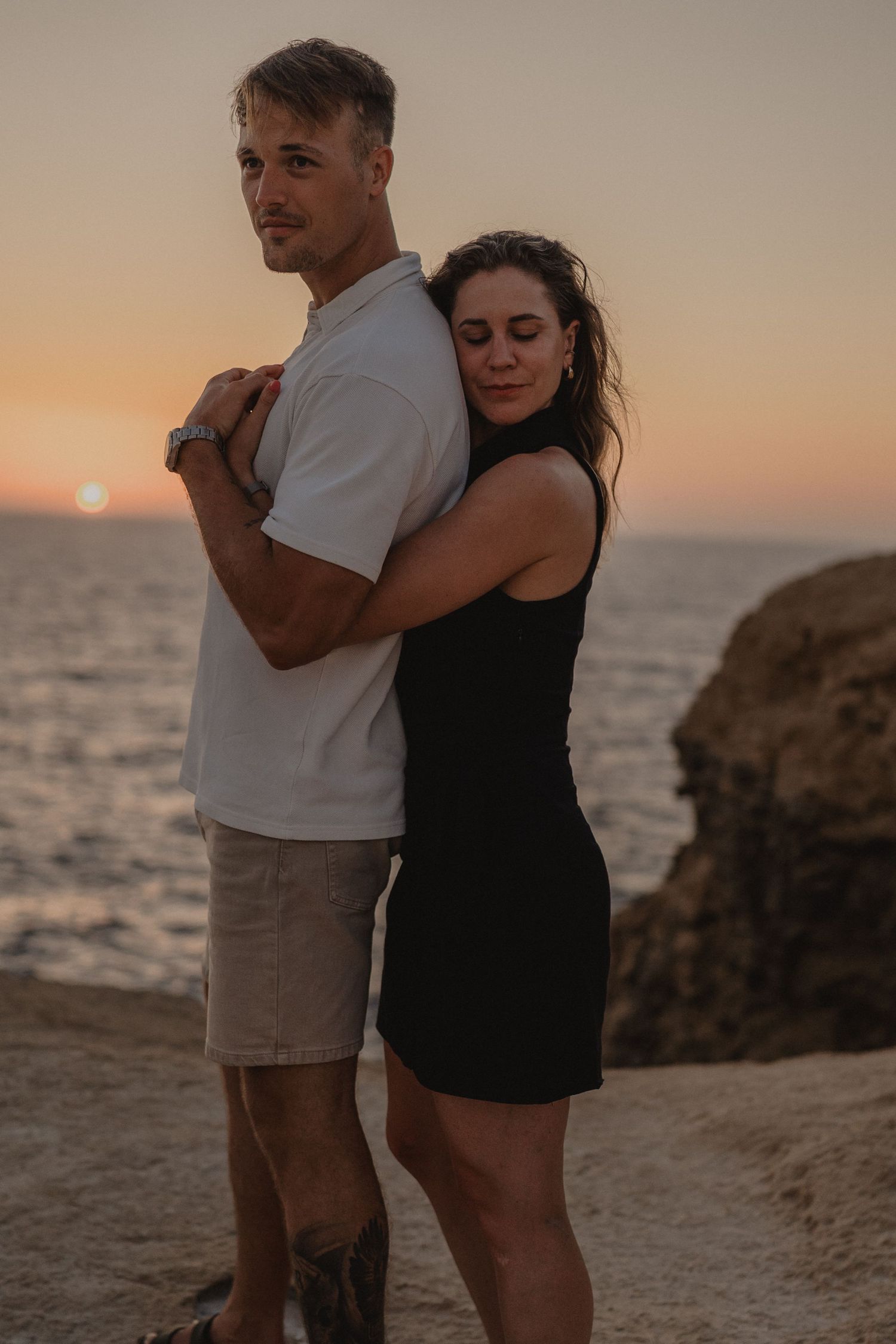 Couple embracing at sunset by the ocean, man in white polo and shorts, woman in black dress, rocky coastline backdrop.