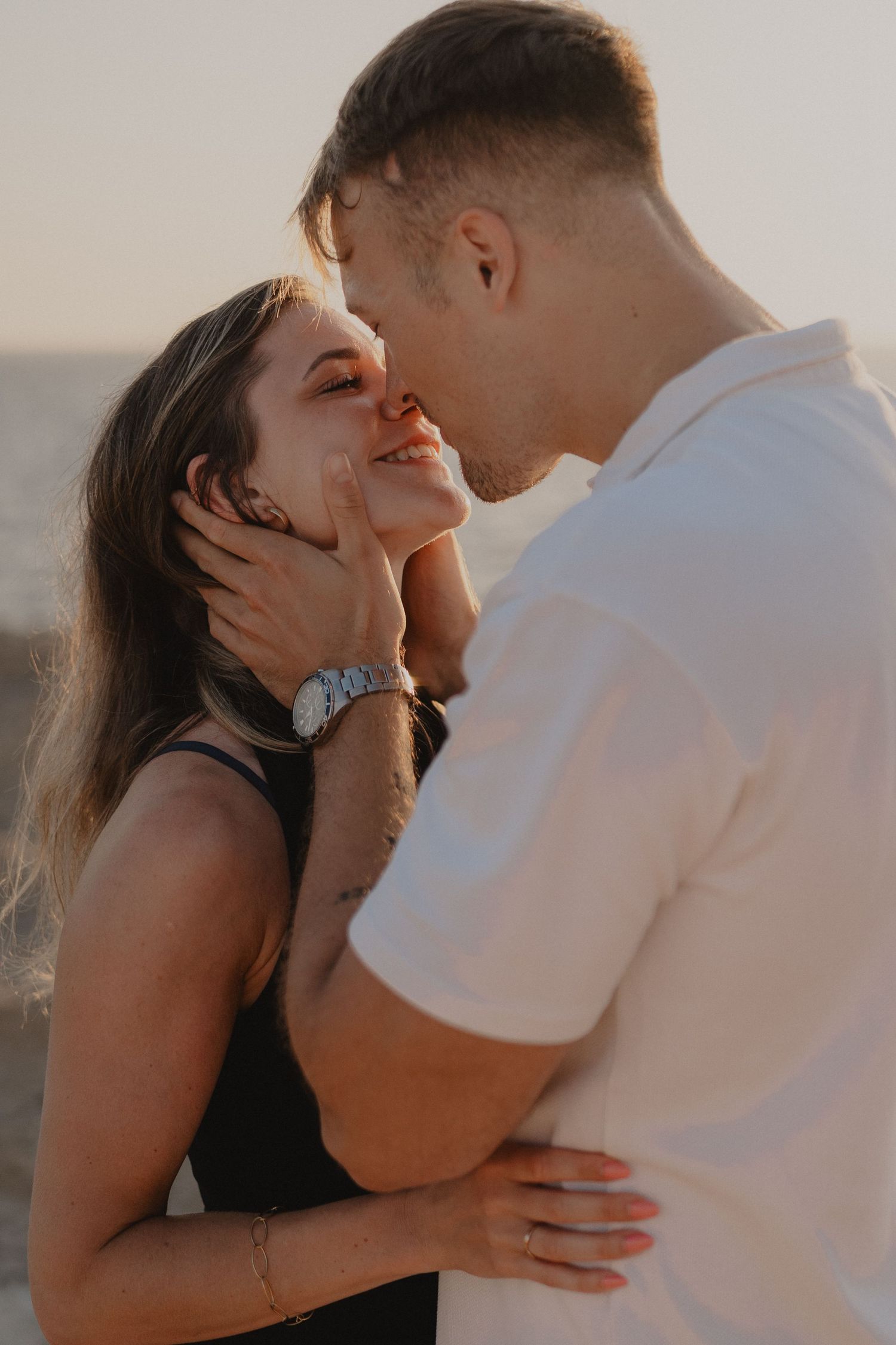 A smiling couple sharing a tender moment at sunset on a beach, man gently cupping womans face as they kiss.