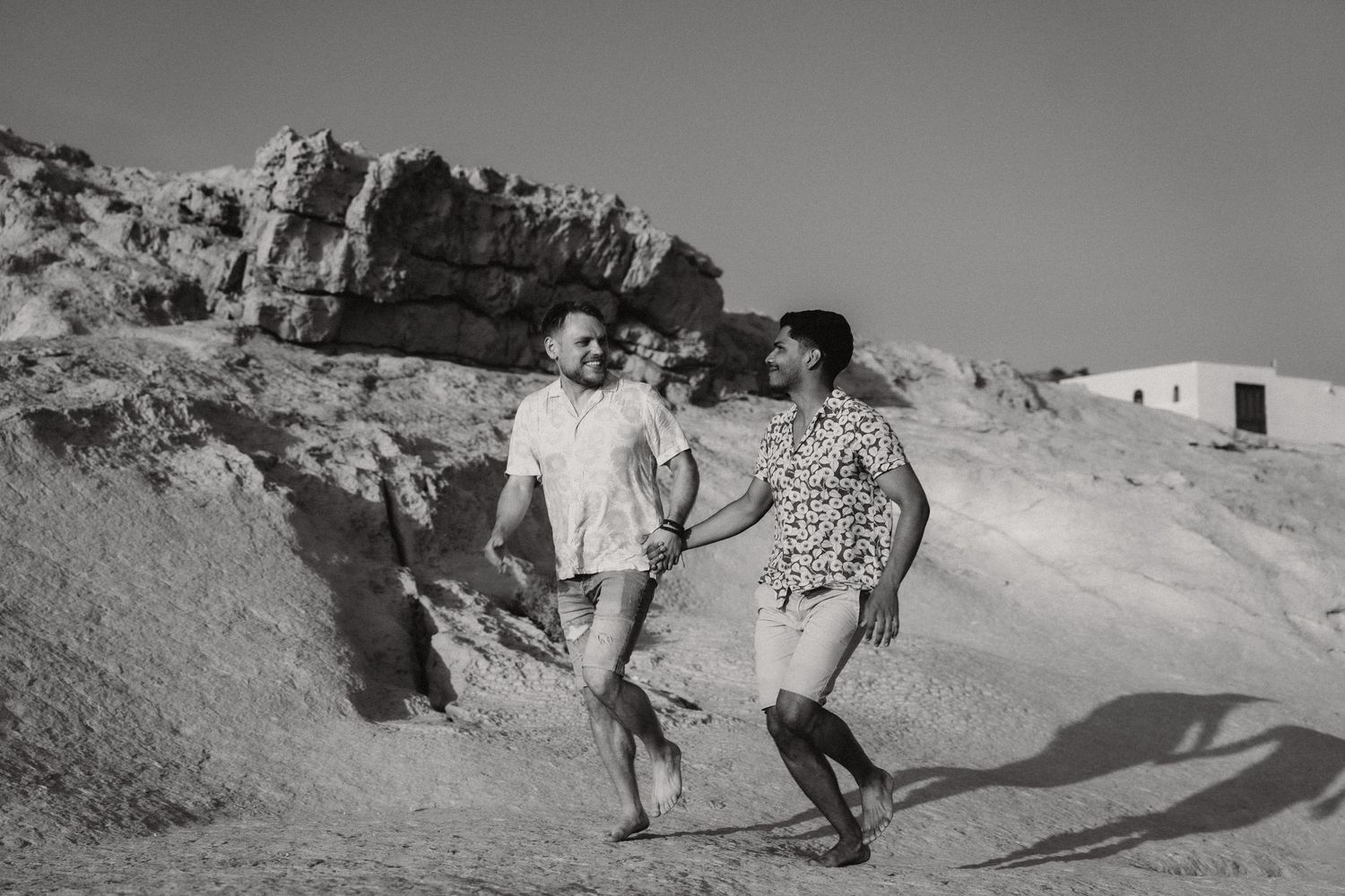Black and white photo of a couple holding hands and walking on a sandy rocky landscape under a clear sky.