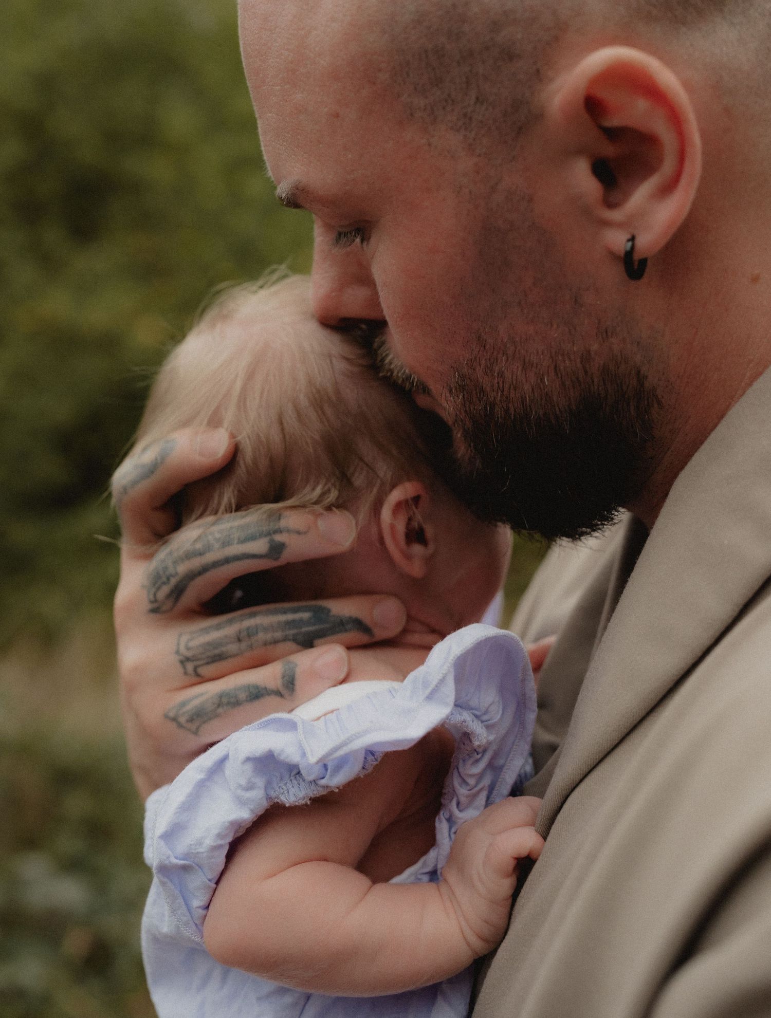 Tattooed father tenderly kissing his baby's head while cradling the infant outdoors.