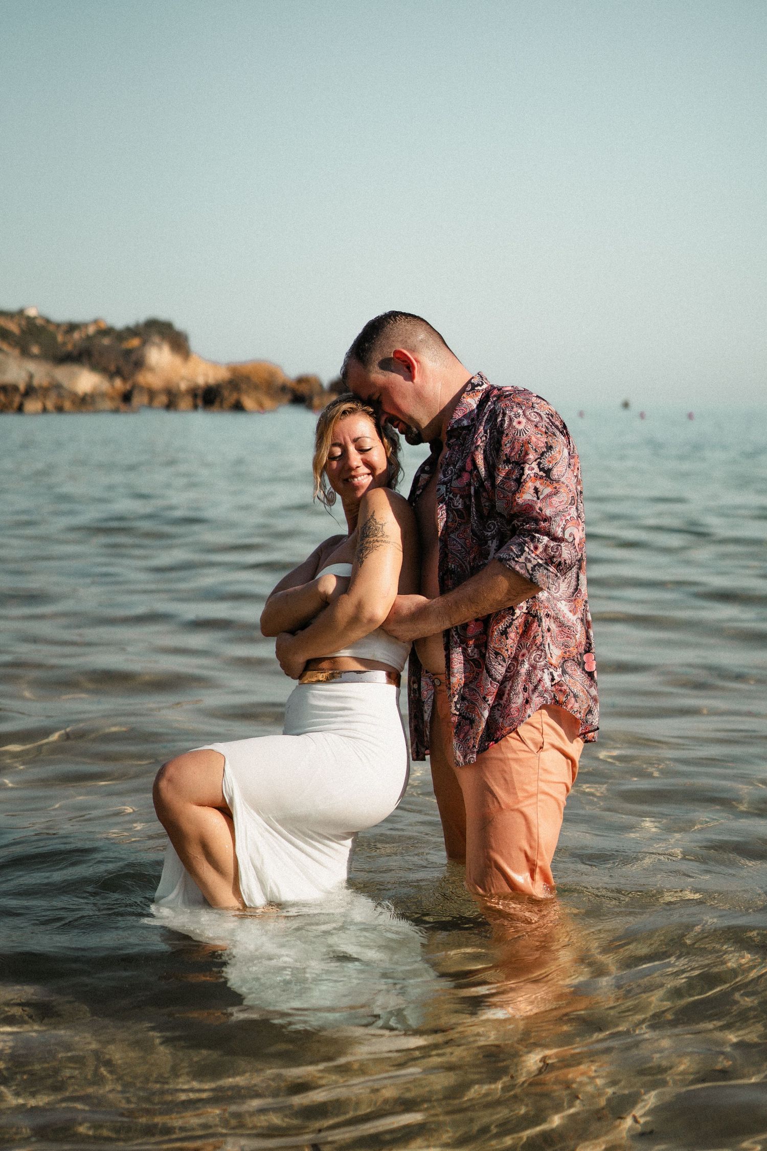 Couple shares playful moments in shallow ocean waters during sunset photoshoot.