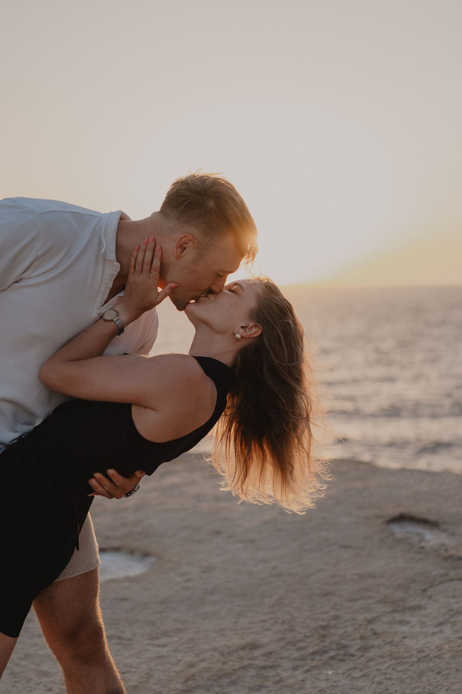 Couple sharing a romantic kiss at sunset by the ocean, woman in black dress dipping back, golden light behind them.