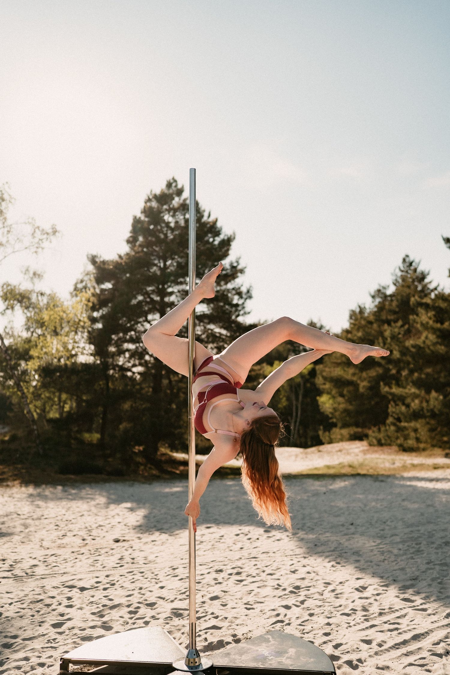 Woman performing an inverted pole dance move outdoors on sandy ground with pine trees in background.