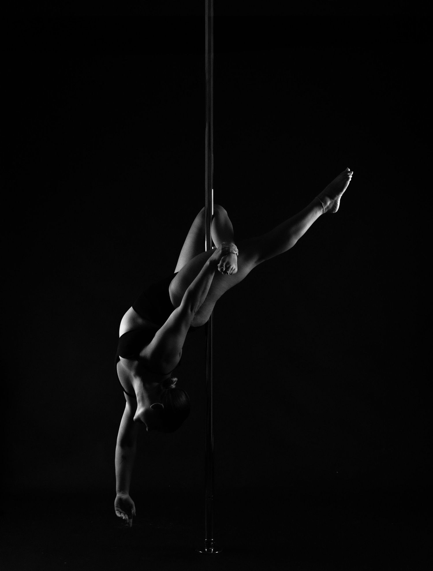 Black and white photo of an athletic pole dancer performing an inverted split pose on a vertical pole against dark background.