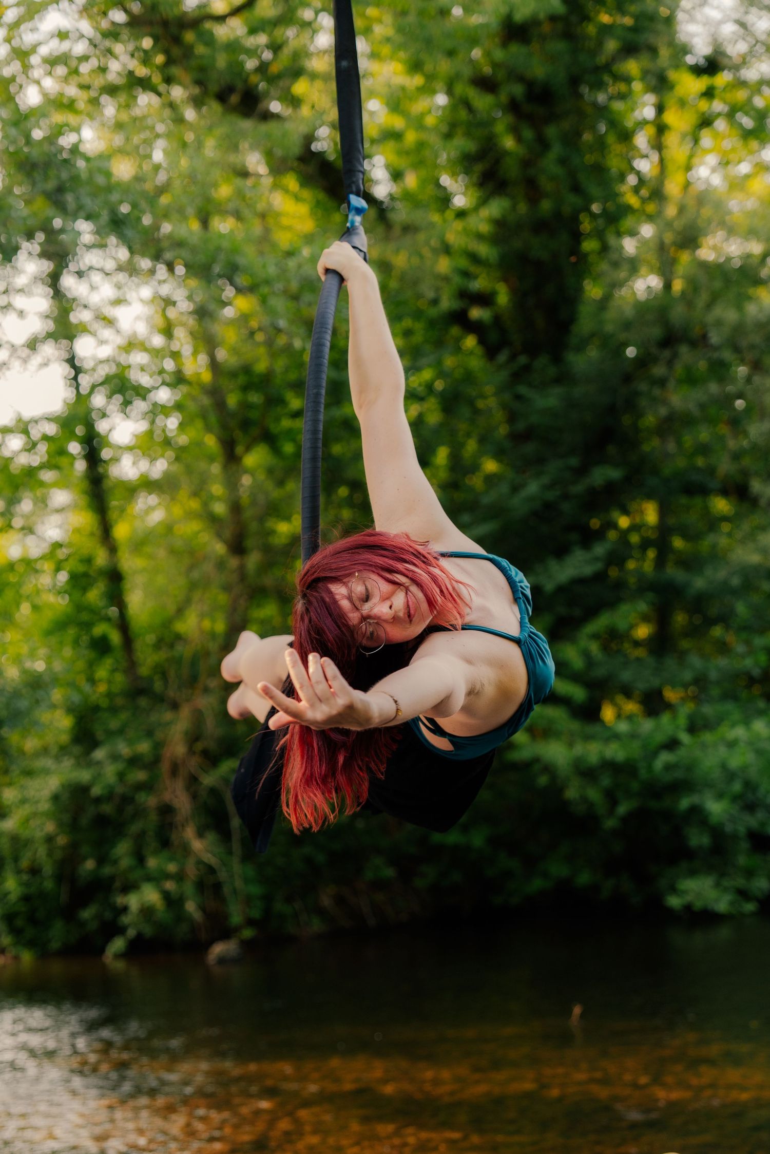 Red-haired woman performing aerial acrobatics on silk straps outdoors near a river surrounded by lush green trees.
