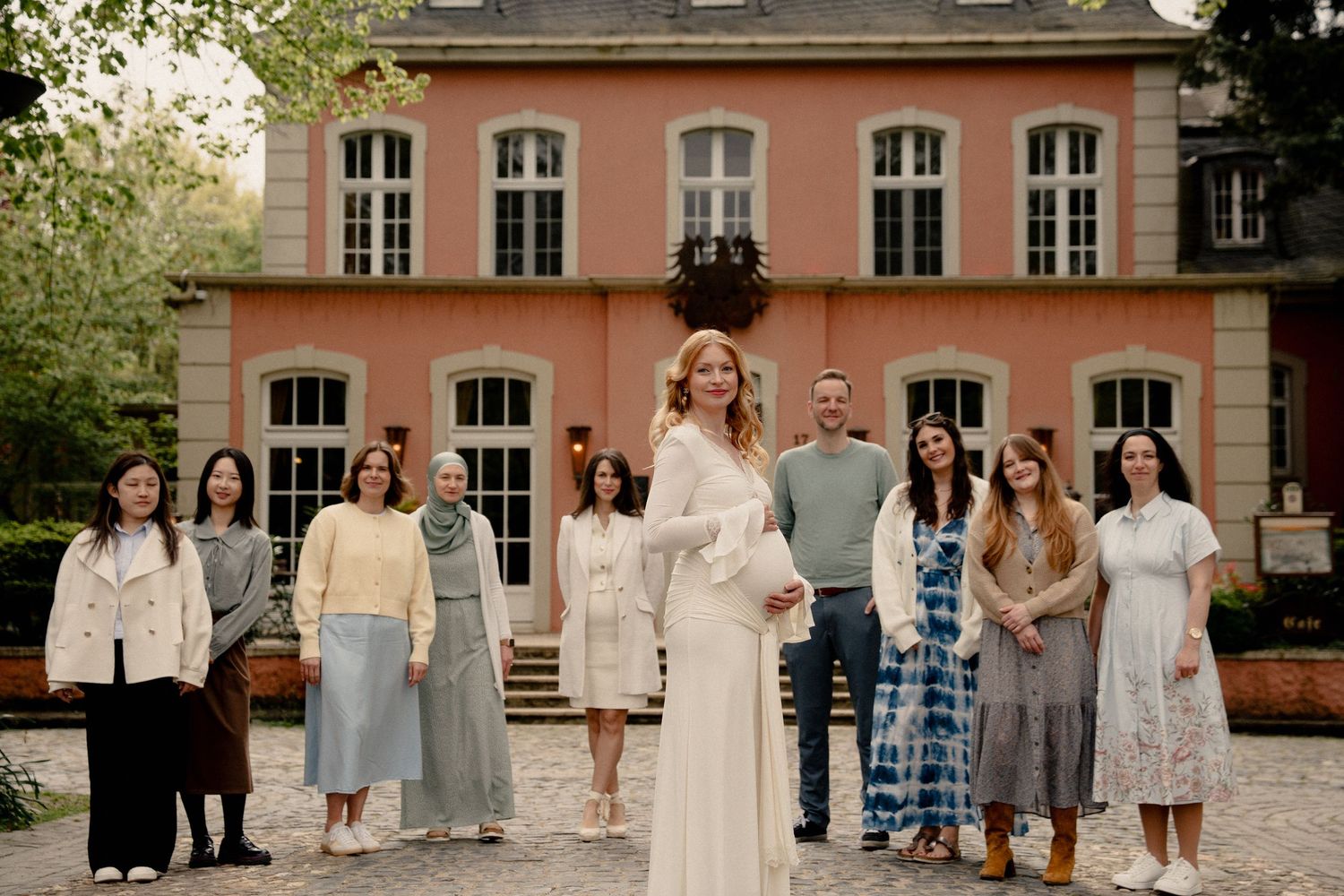 Elegant group portrait of people in light attire standing before a pink historic villa.