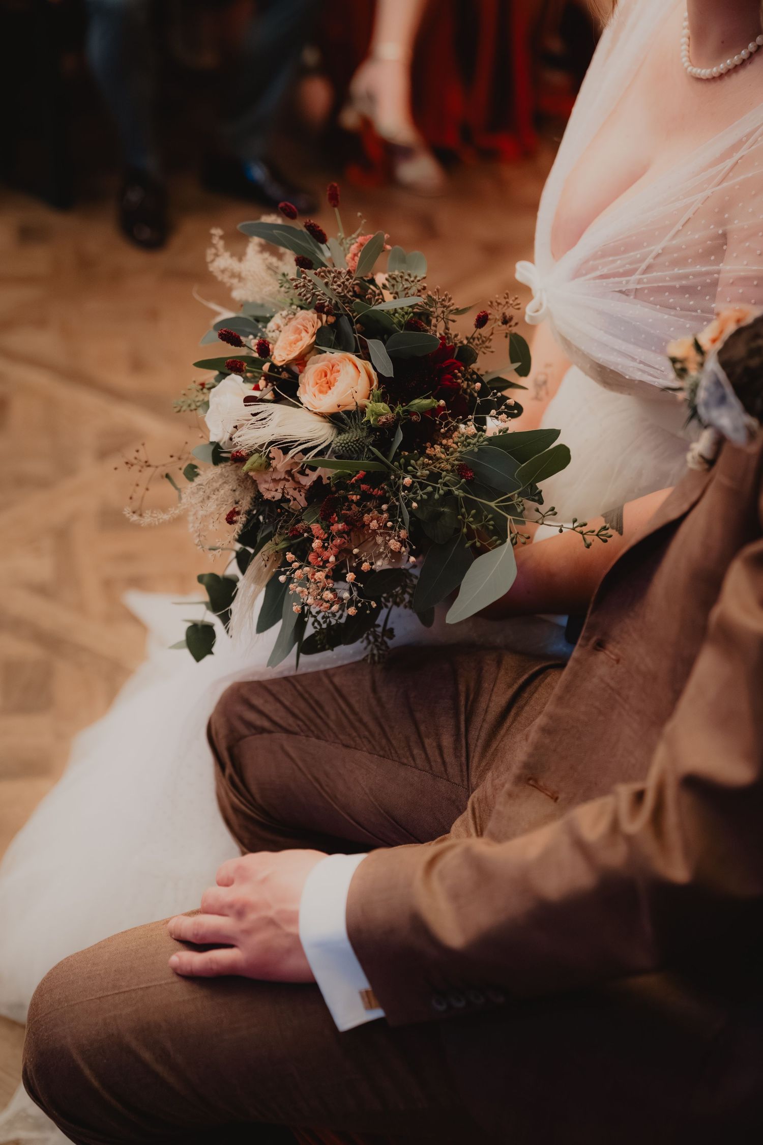 Wedding bouquet with peach roses and greenery resting on brown suit fabric.