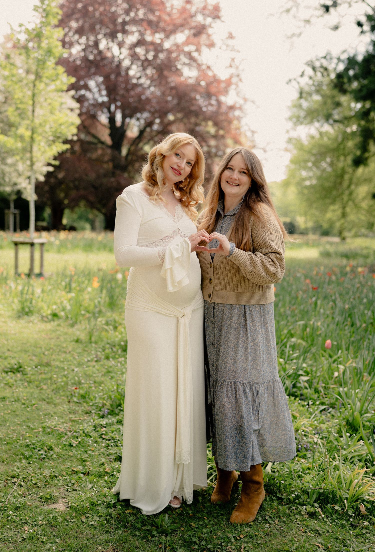 Pregnant woman in white dress posing with friend in spring garden with colorful flowers.
