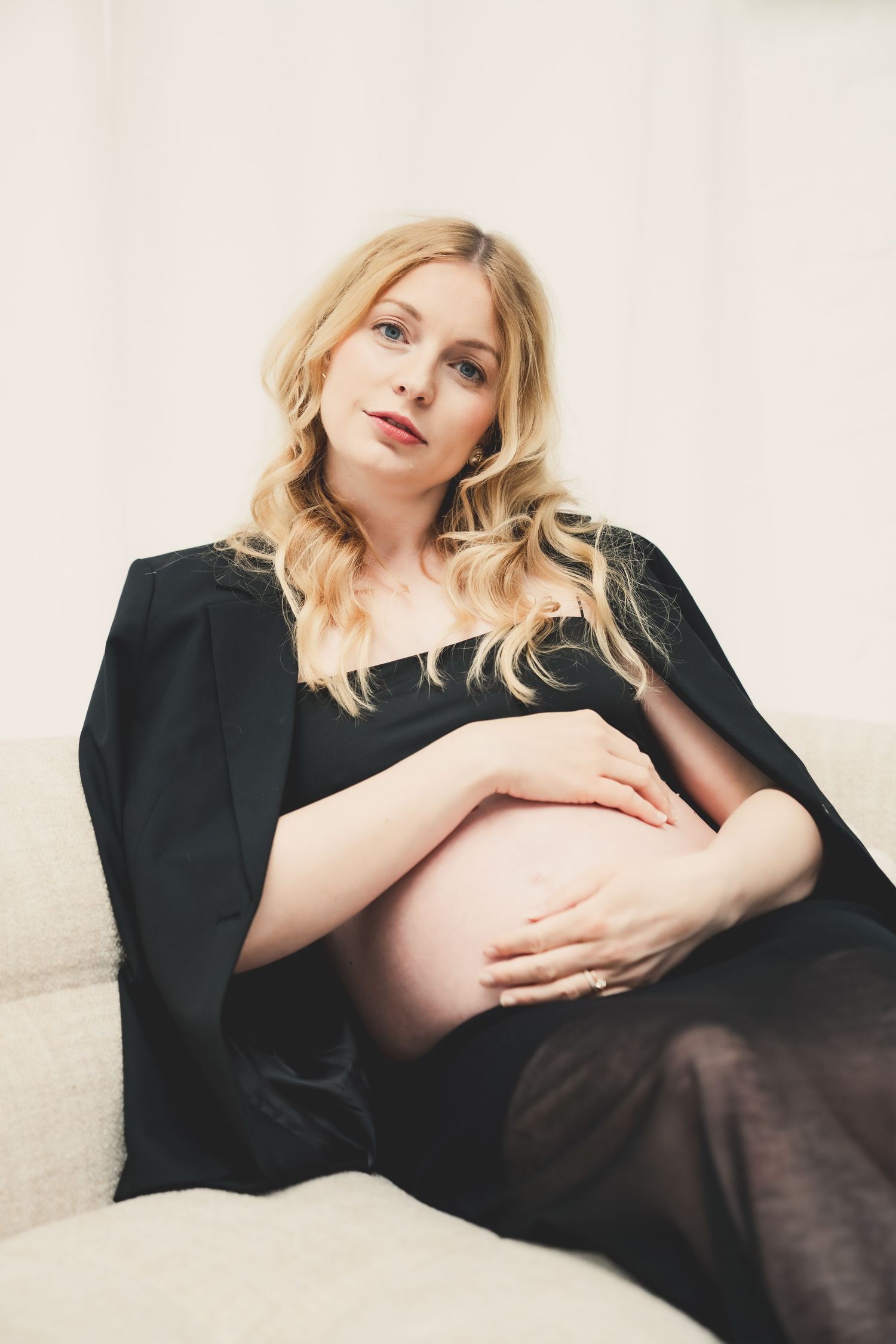 Pregnant woman in black robe sitting on sofa, cradling her baby bump with both hands, looking at camera.