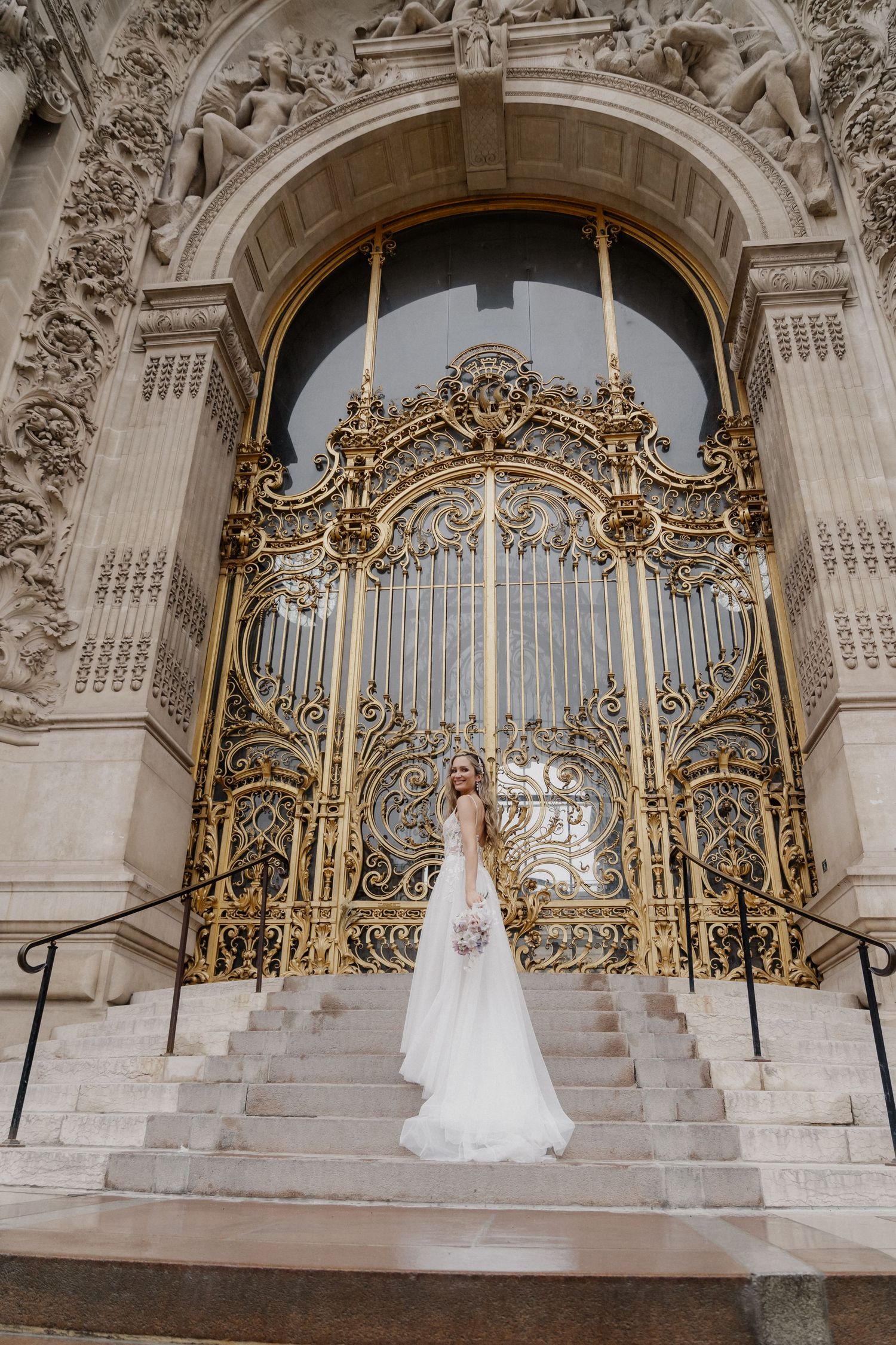 A bride in a white gown stands before ornate golden gates at the entrance of a grand stone building with baroque architecture.