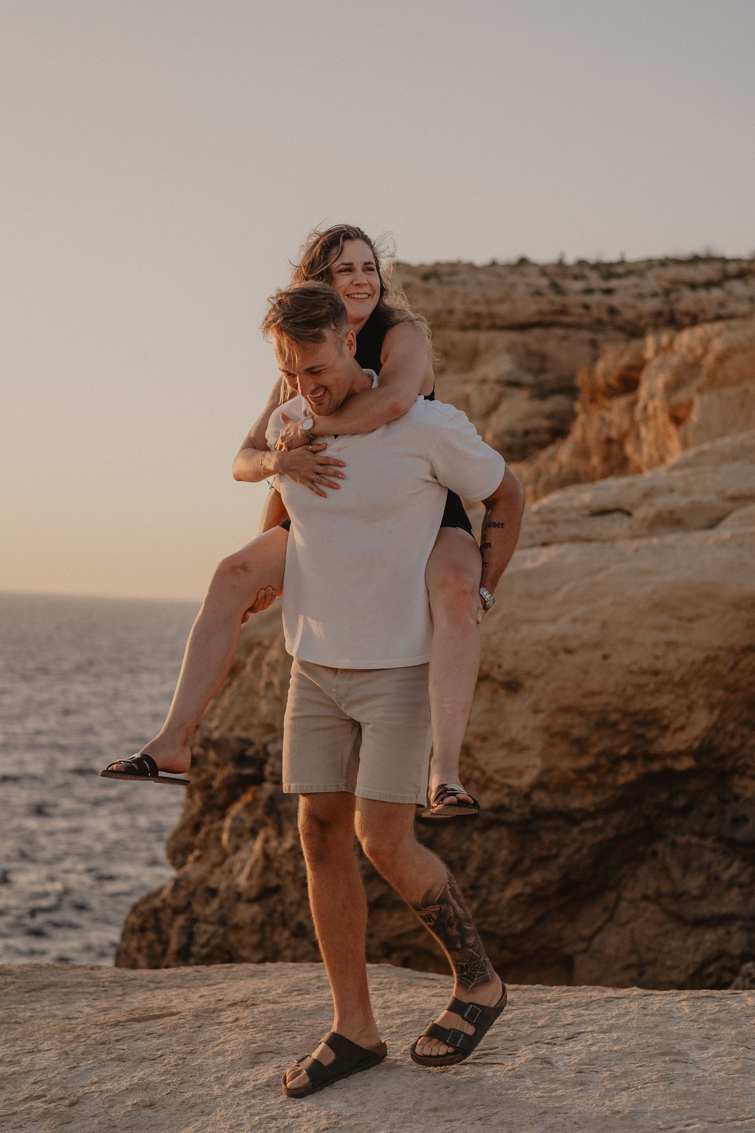 A man giving a woman a piggyback ride on rocky coastal cliffs at sunset, both smiling and enjoying the moment.