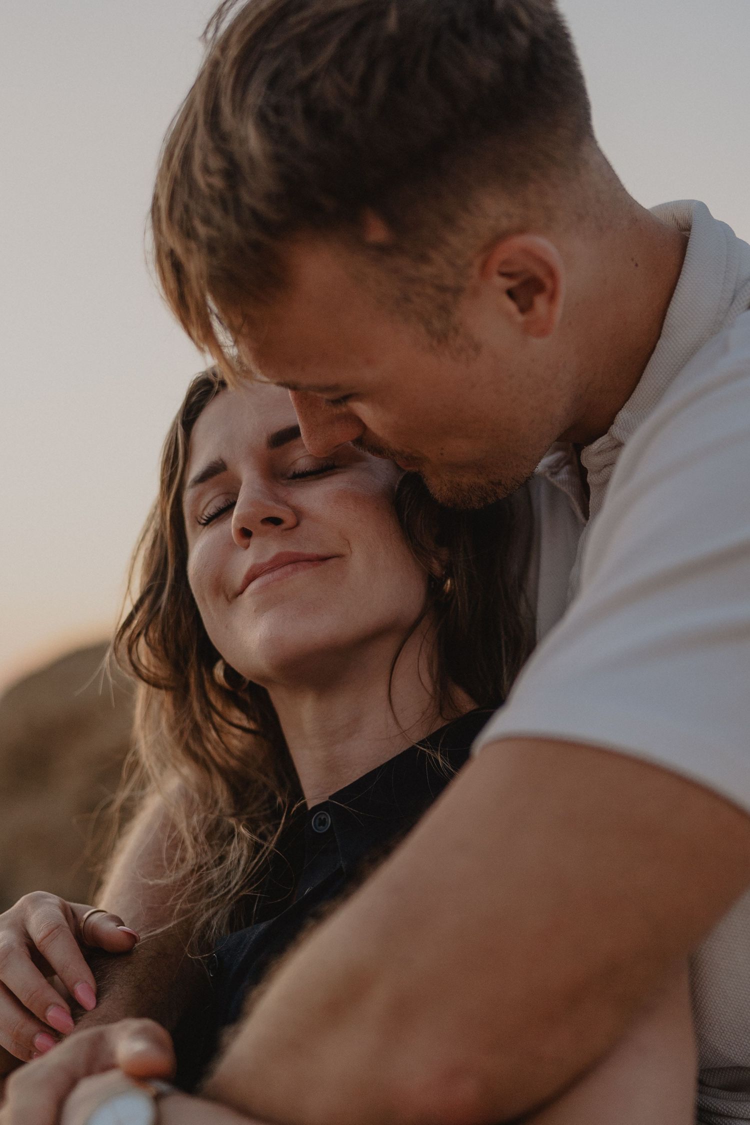 A couple shares a tender embrace at sunset, with the man kissing the smiling woman's forehead.