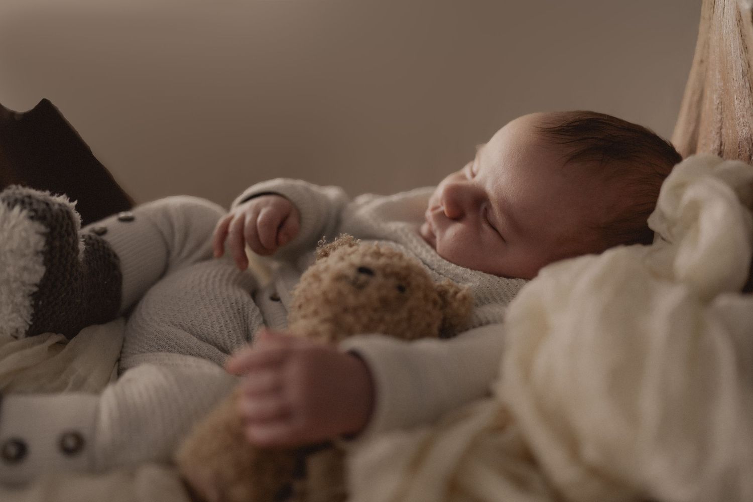 An infant rests peacefully on cream-colored bedding with stuffed animals nearby.