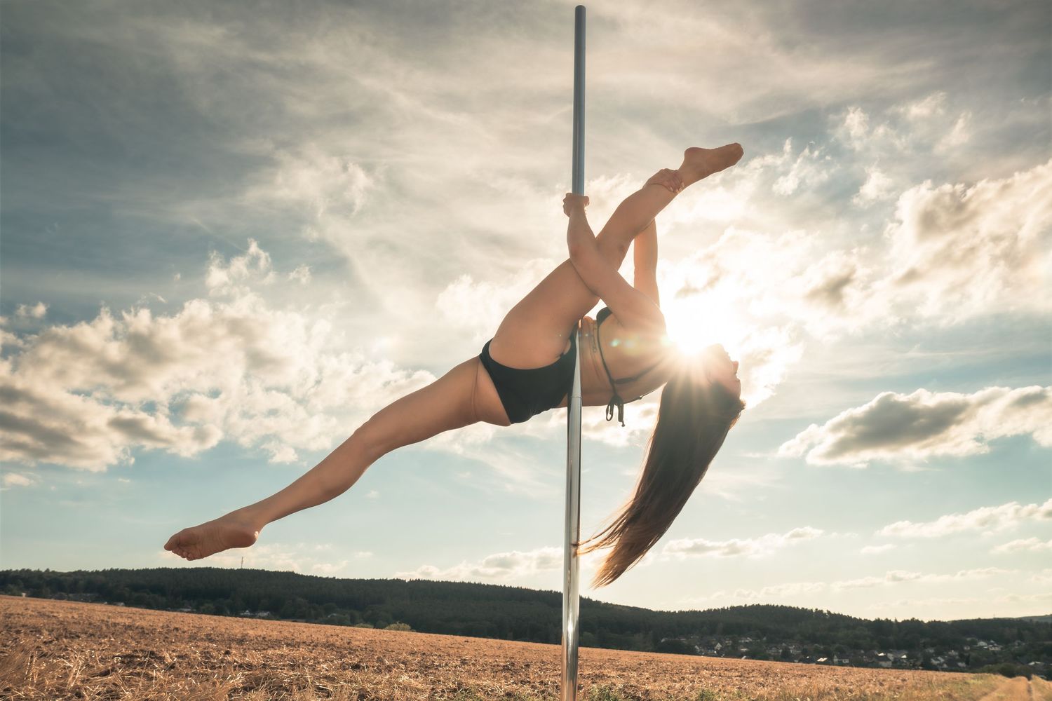 Woman performing aerial pole dance outdoors against a dramatic cloudy sky in a sunlit field.