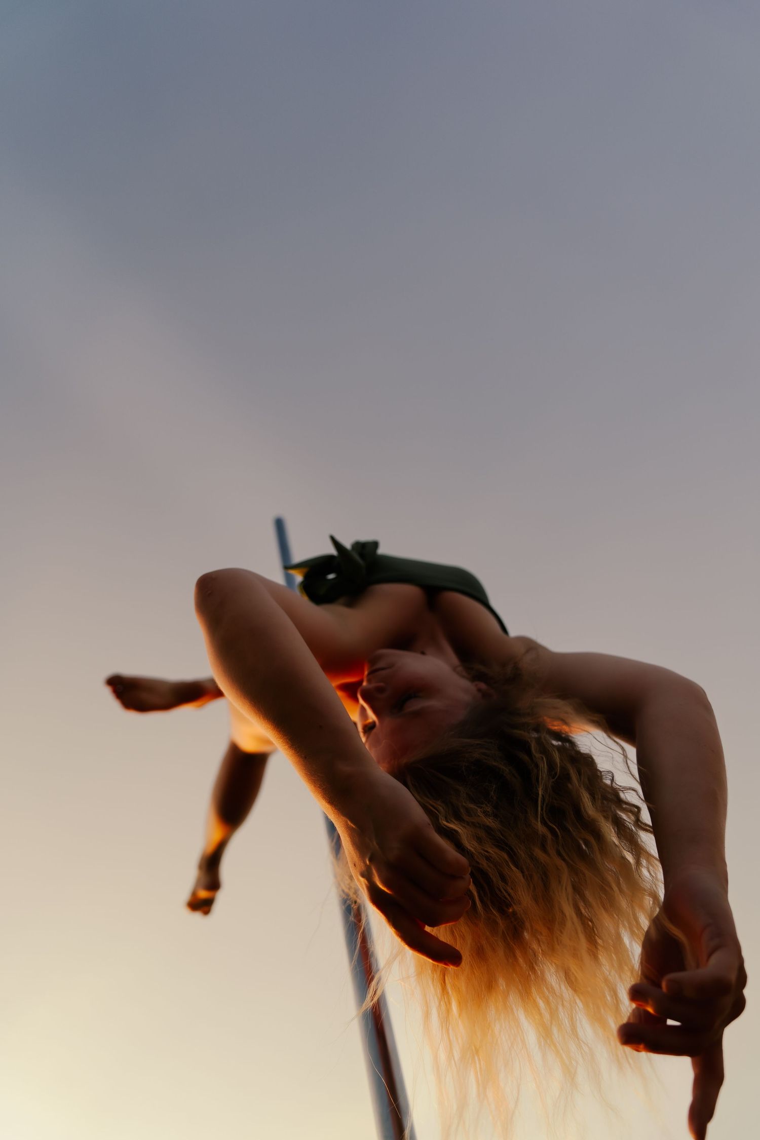 A flexible female acrobat bends backward on a pole against a dramatic sunset sky, her long blonde hair flowing downward.