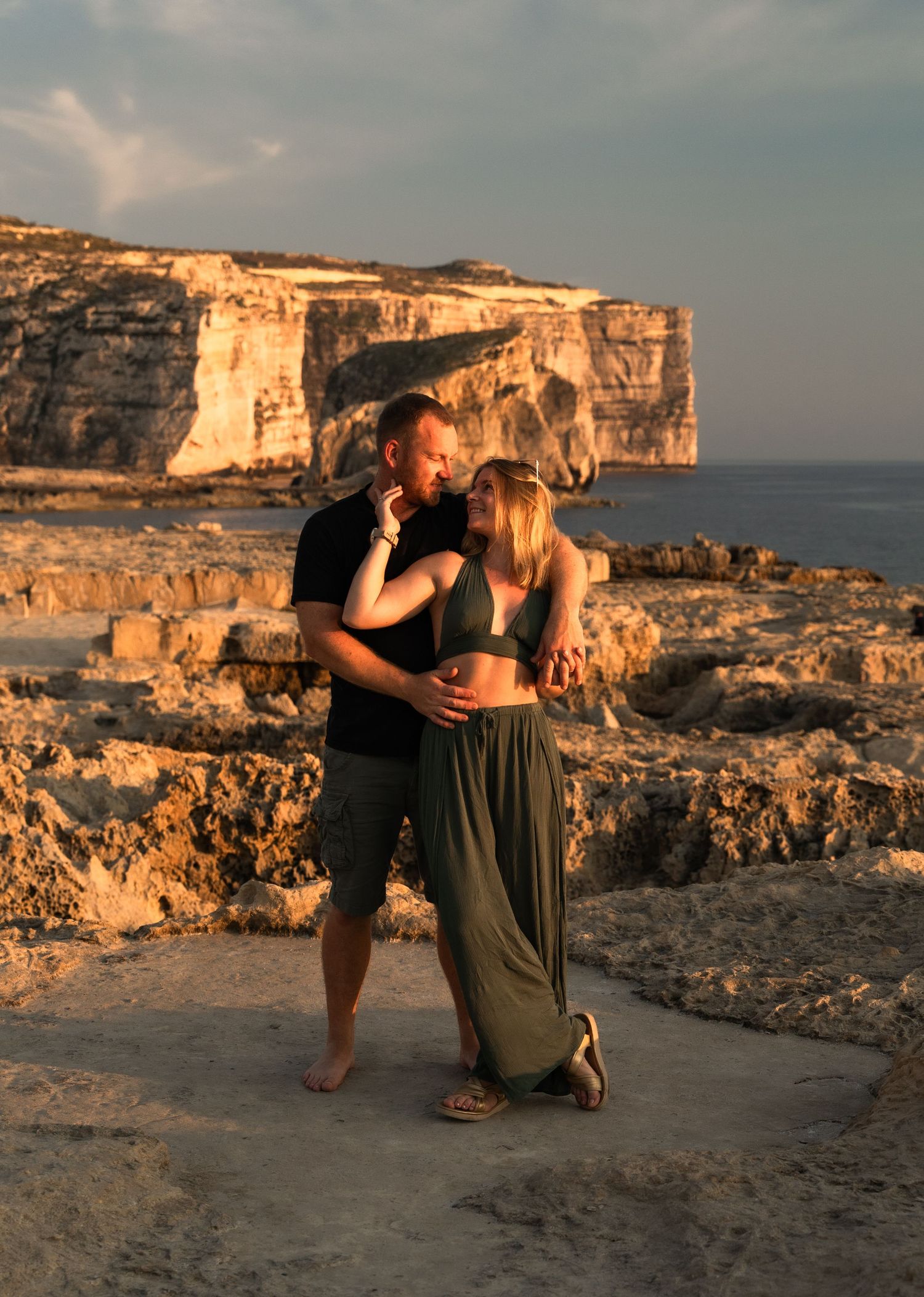 Couple embracing at sunset on rocky coastal cliffs with turquoise sea and limestone formations in background.