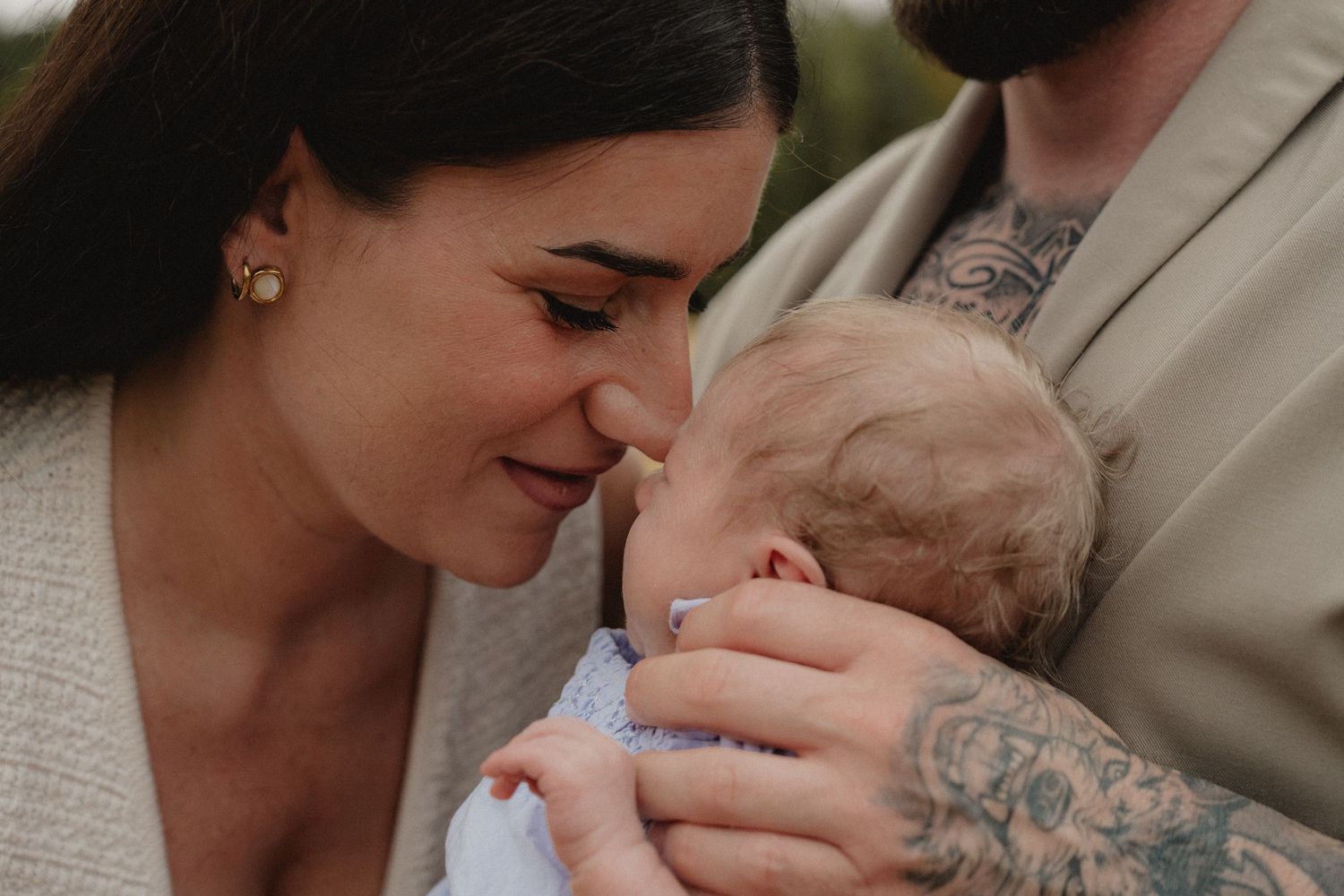 A mother tenderly nuzzles her newborn baby held by a tattooed partner outdoors in soft natural light.