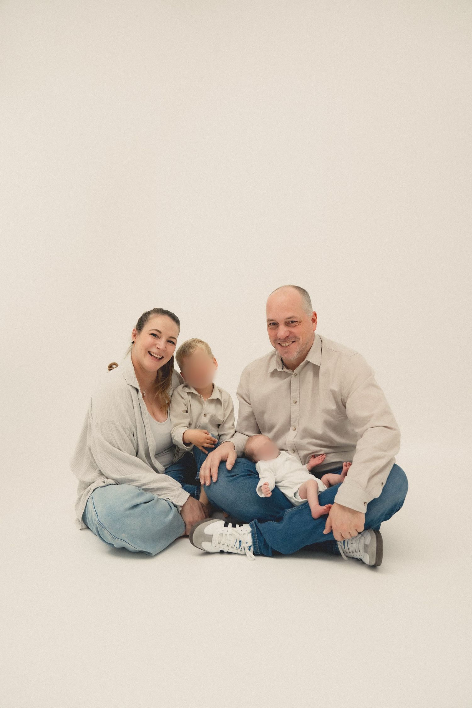 Happy family of three sitting on white studio floor, parents smiling with toddler and newborn baby.